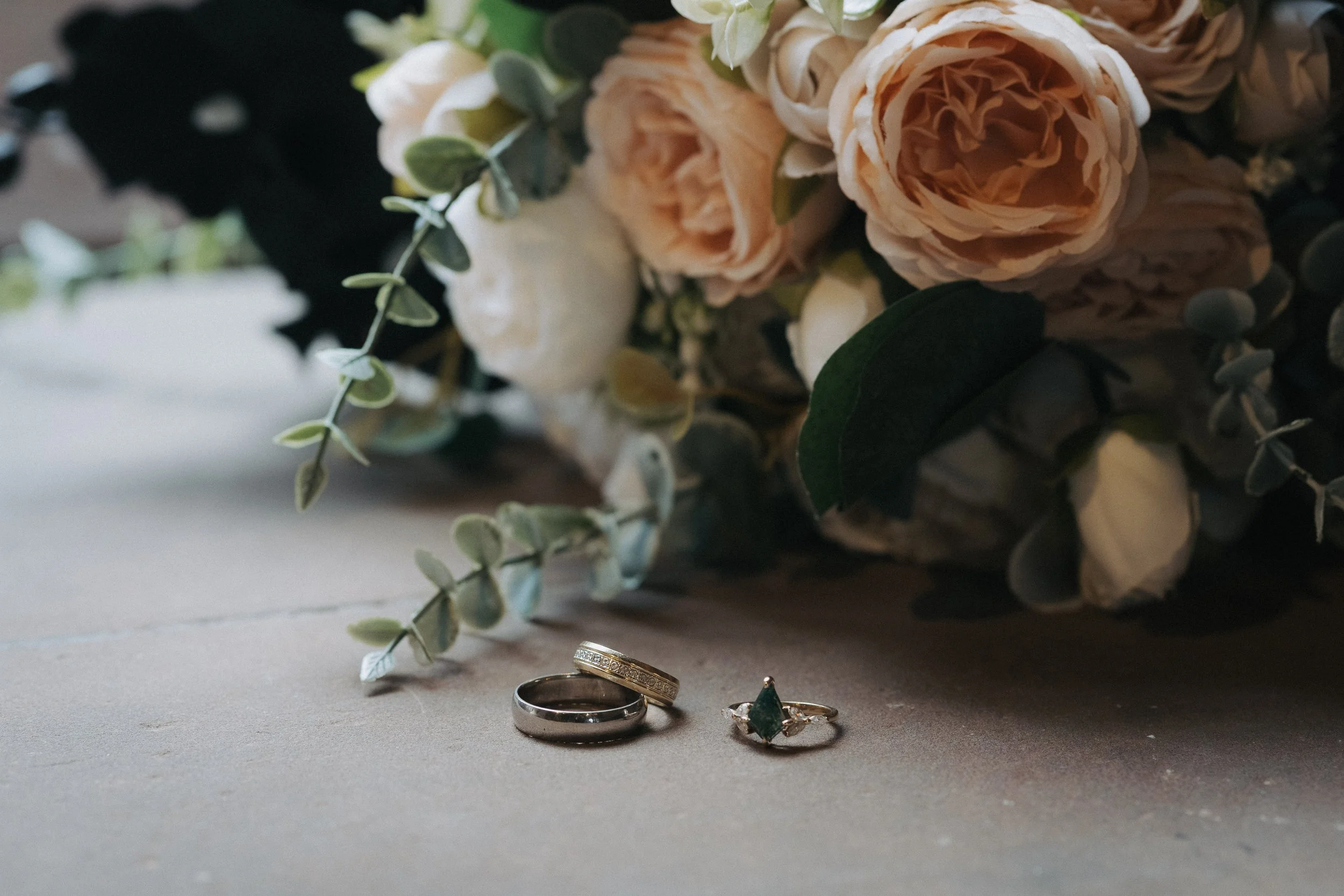 Three wedding rings placed on a gray surface with a bouquet of soft pink roses, white flowers, and green leaves in the background. - captured by an Edinburgh wedding photographer