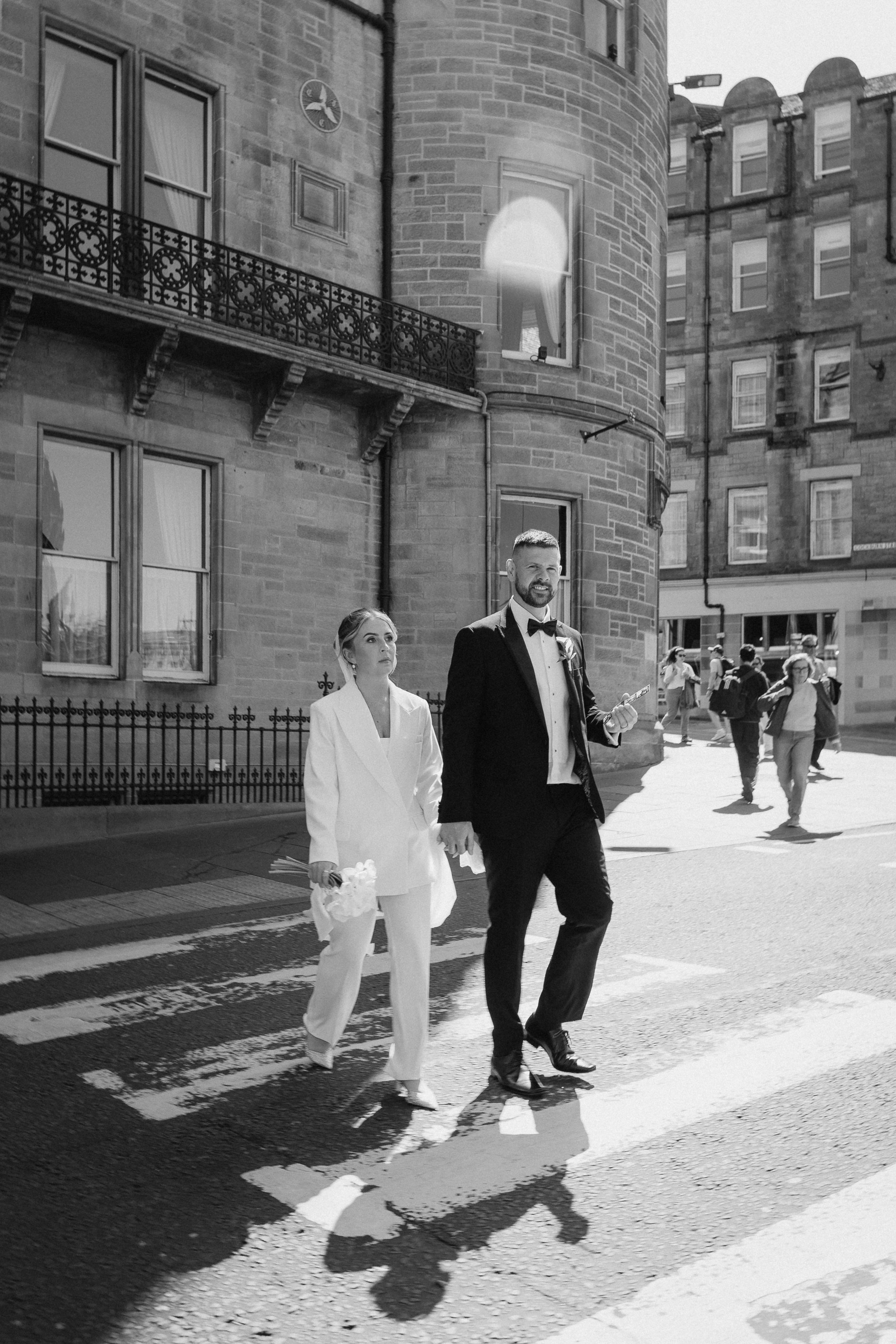 A black and white photo of a couple dressed in formal attire crossing a street in an urban area, holding hands. The woman is wearing a white suit and holding flowers, and the man is in a black tuxedo with a bow tie, smiling. Pedestrians are walking i