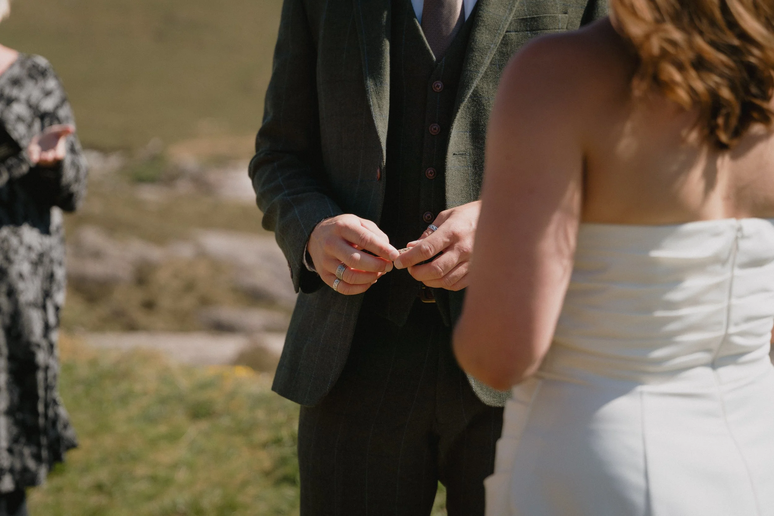 A couple during a wedding ceremony exchanging rings outdoors, with the groom wearing a dark plaid suit and the bride in a strapless white wedding dress. - captured by an Edinburgh wedding photographer