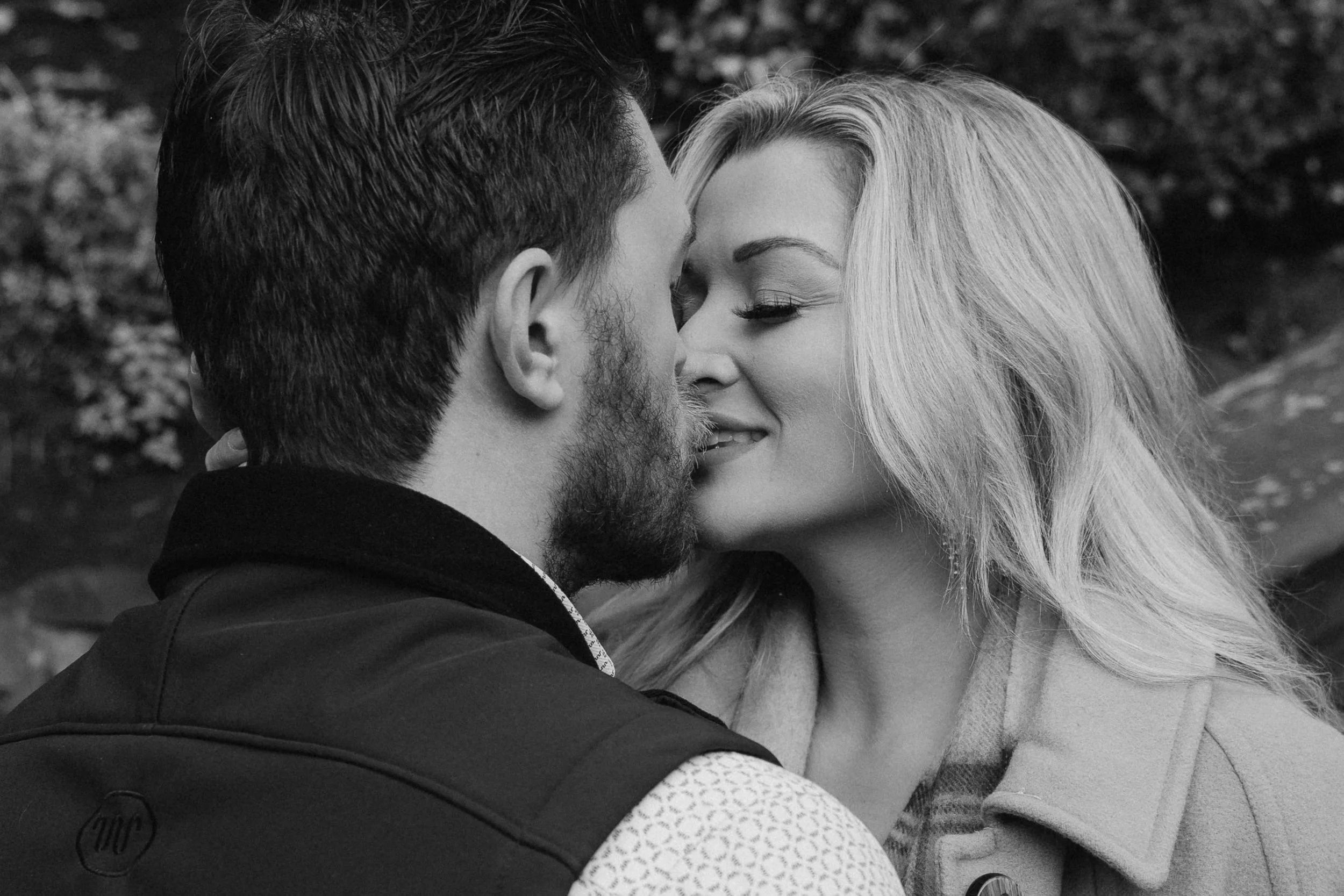 A black-and-white photo of a couple close together, about to kiss, with outdoor trees in the background. - captured by an Edinburgh wedding photographer