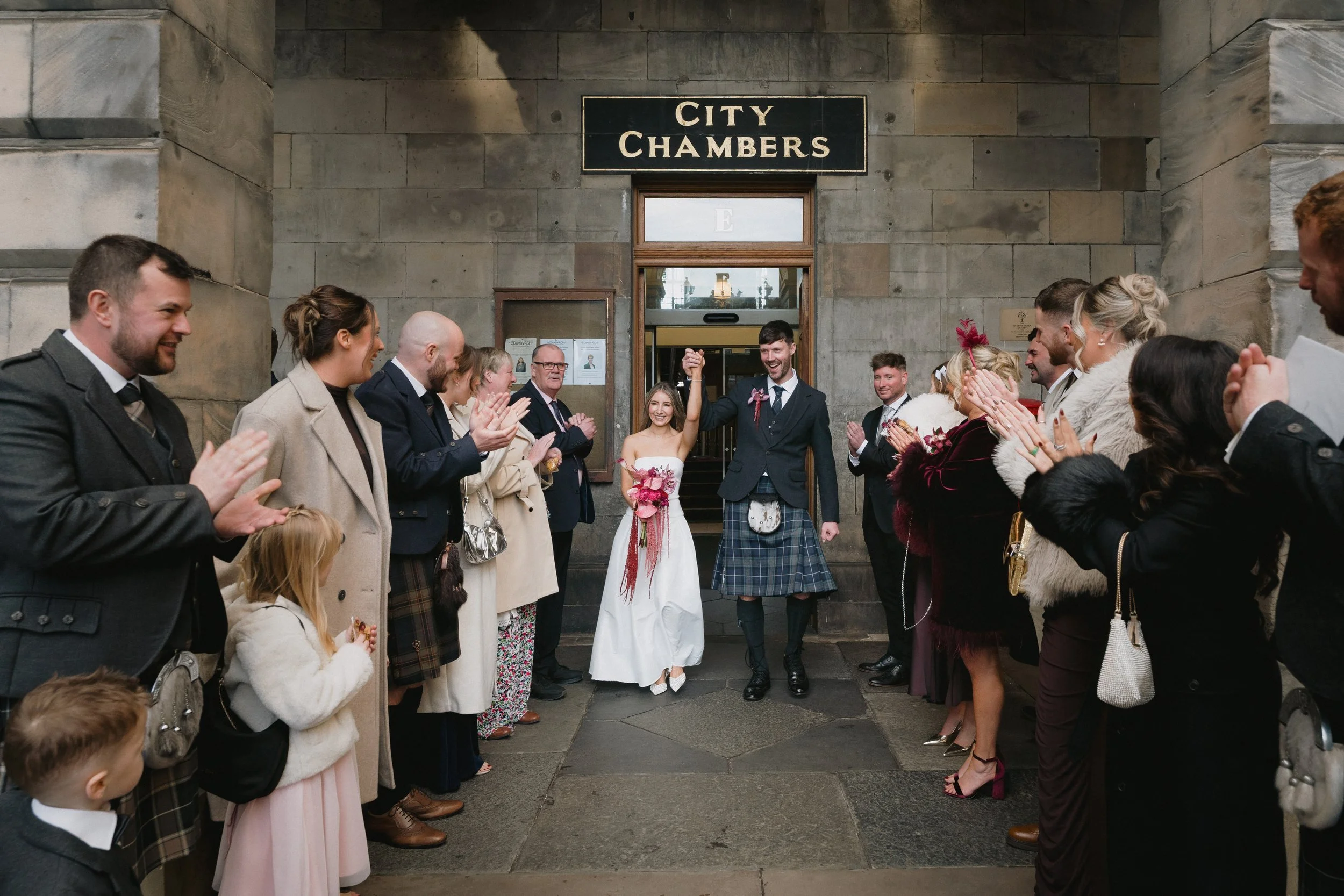Newlywed couple holding hands and celebrating outside City Chambers with family and friends clapping and cheering. - captured by an Edinburgh wedding photographer