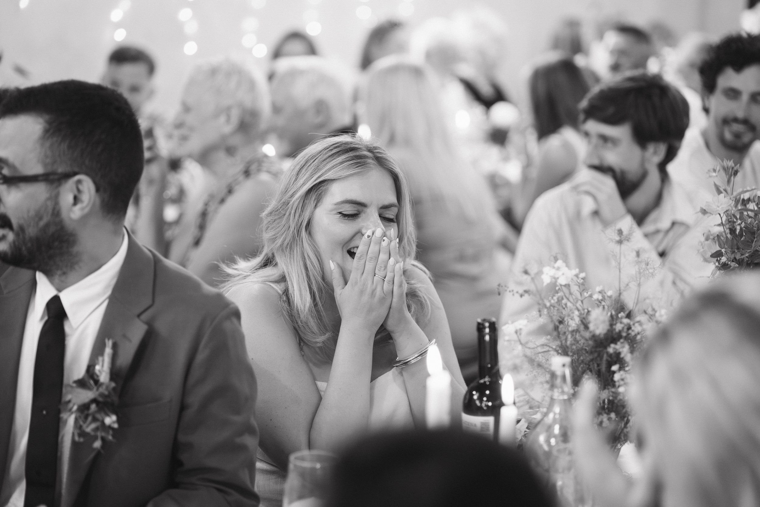 A woman at a wedding reception is smiling with her eyes closed and her hands covering her mouth, appearing happy and surprised. She is seated at a table with a floral centerpiece and bottles of beer nearby, surrounded by other guests in formal attire