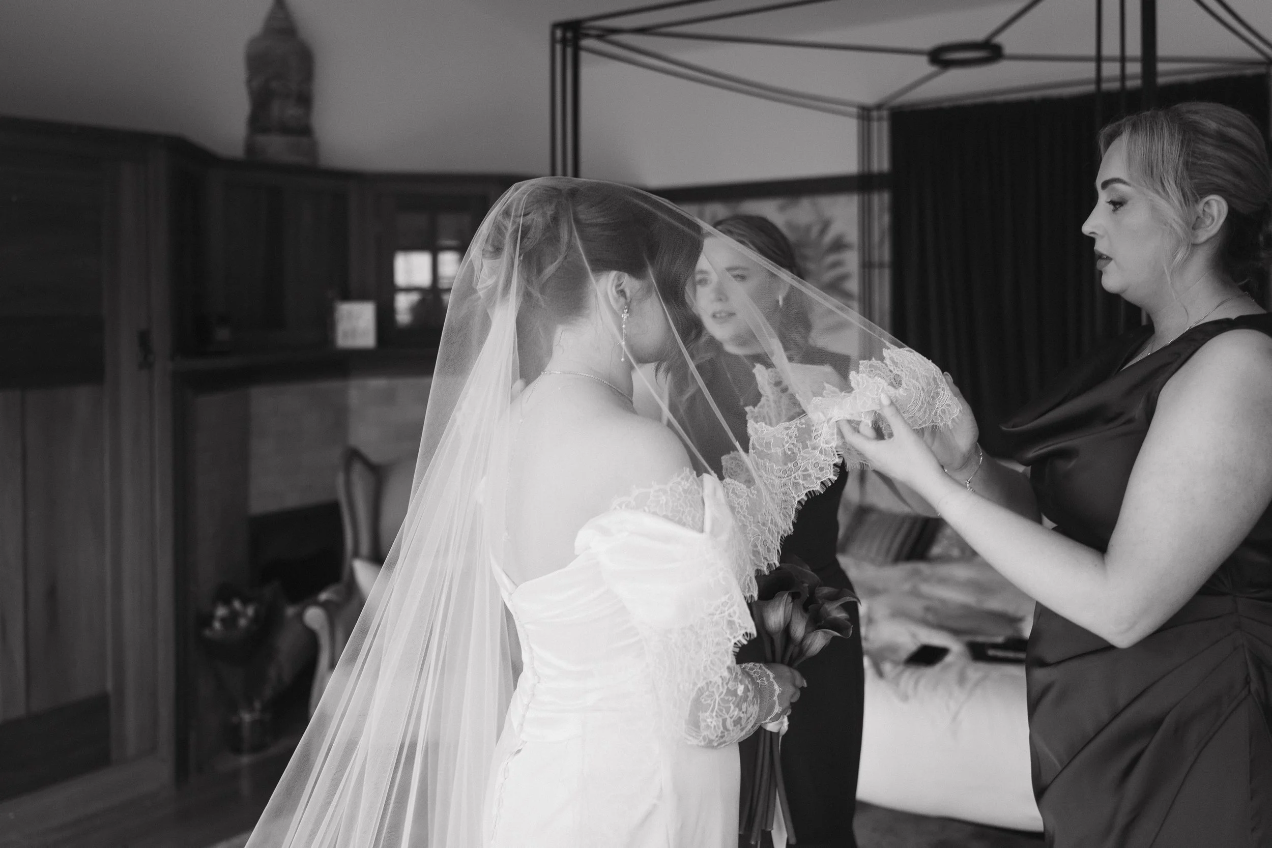 A bride in a wedding dress with a veil is having her headpiece adjusted by a woman in a satin dress while another woman stands nearby watching. - captured by an Edinburgh wedding photographer