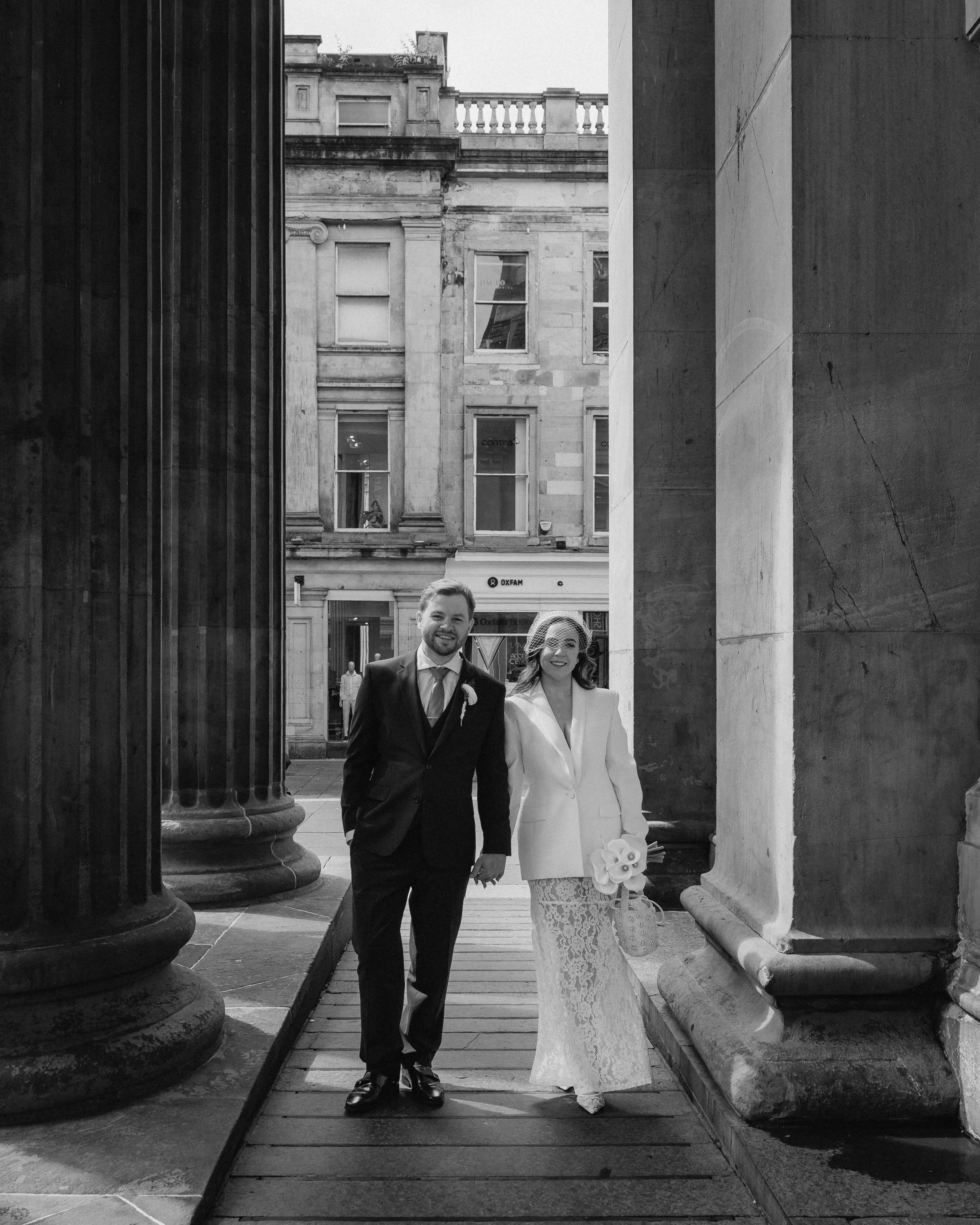 A smiling bride and groom holding hands and walking through a historic building's columns in black and white. - captured by an Edinburgh wedding photographer