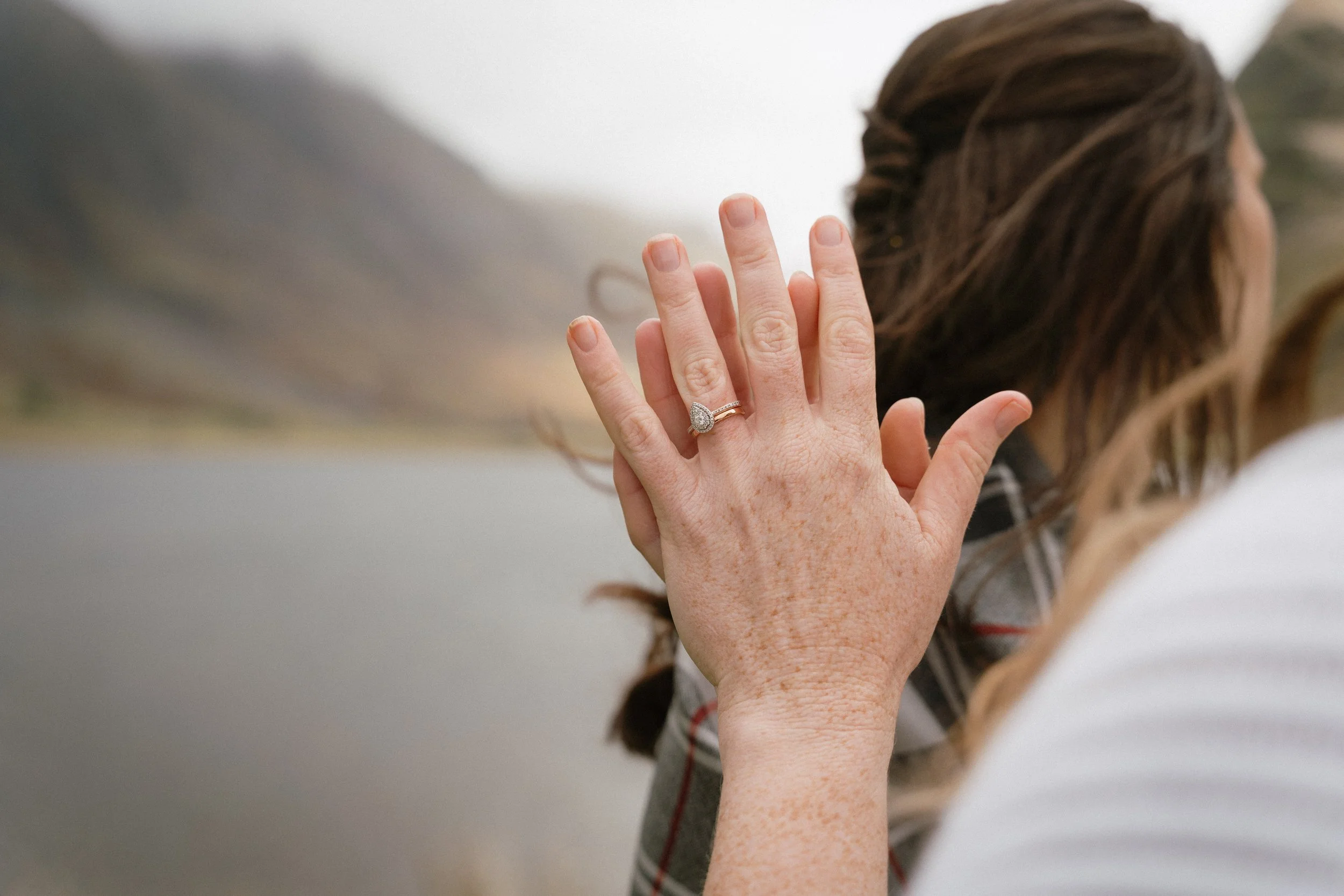 Close-up of a woman's hand with a diamond engagement ring and wedding band, held up near her face with a blurred outdoor background. - captured by an Edinburgh wedding photographer