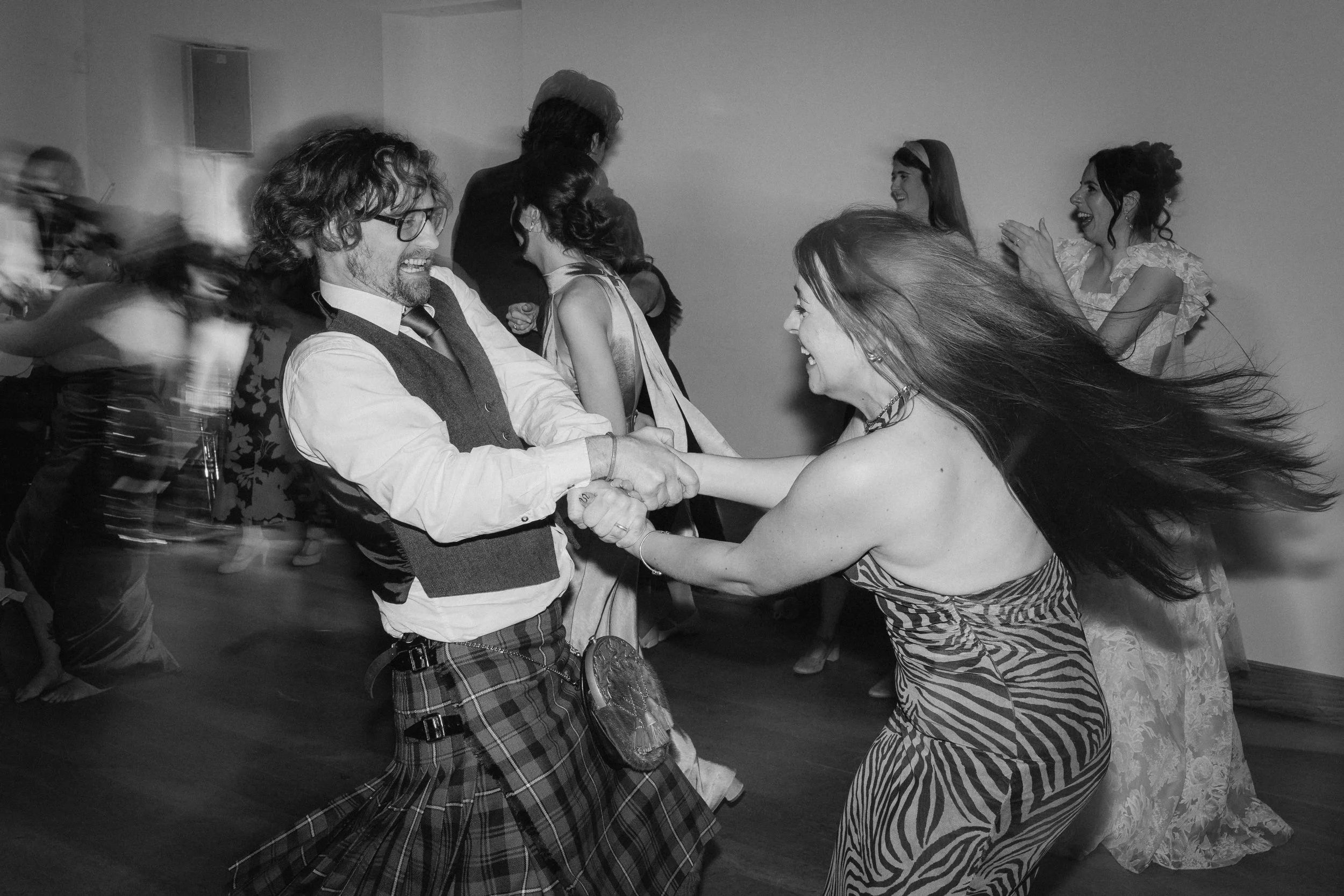 People dancing and having fun at a party, with a man in a kilt and a woman with long hair dancing in the foreground. - captured by an Edinburgh wedding photographer