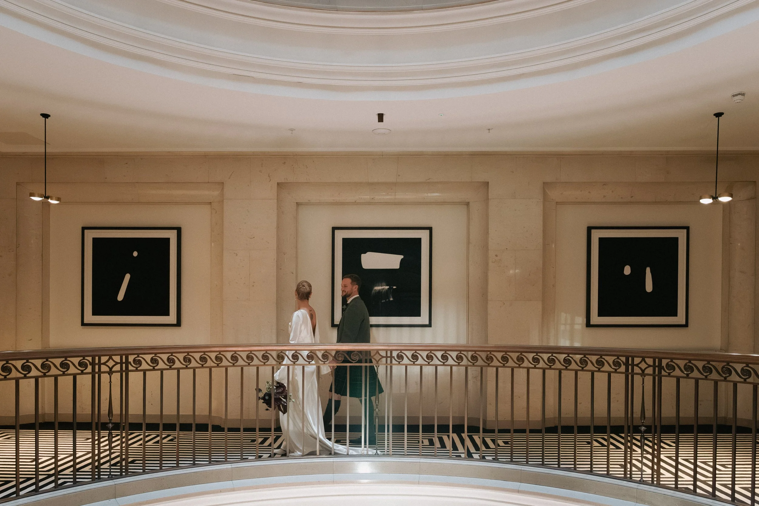 A bride and groom in wedding attire walking on a balcony inside a building with modern abstract art on the walls. - captured by an Edinburgh wedding photographer