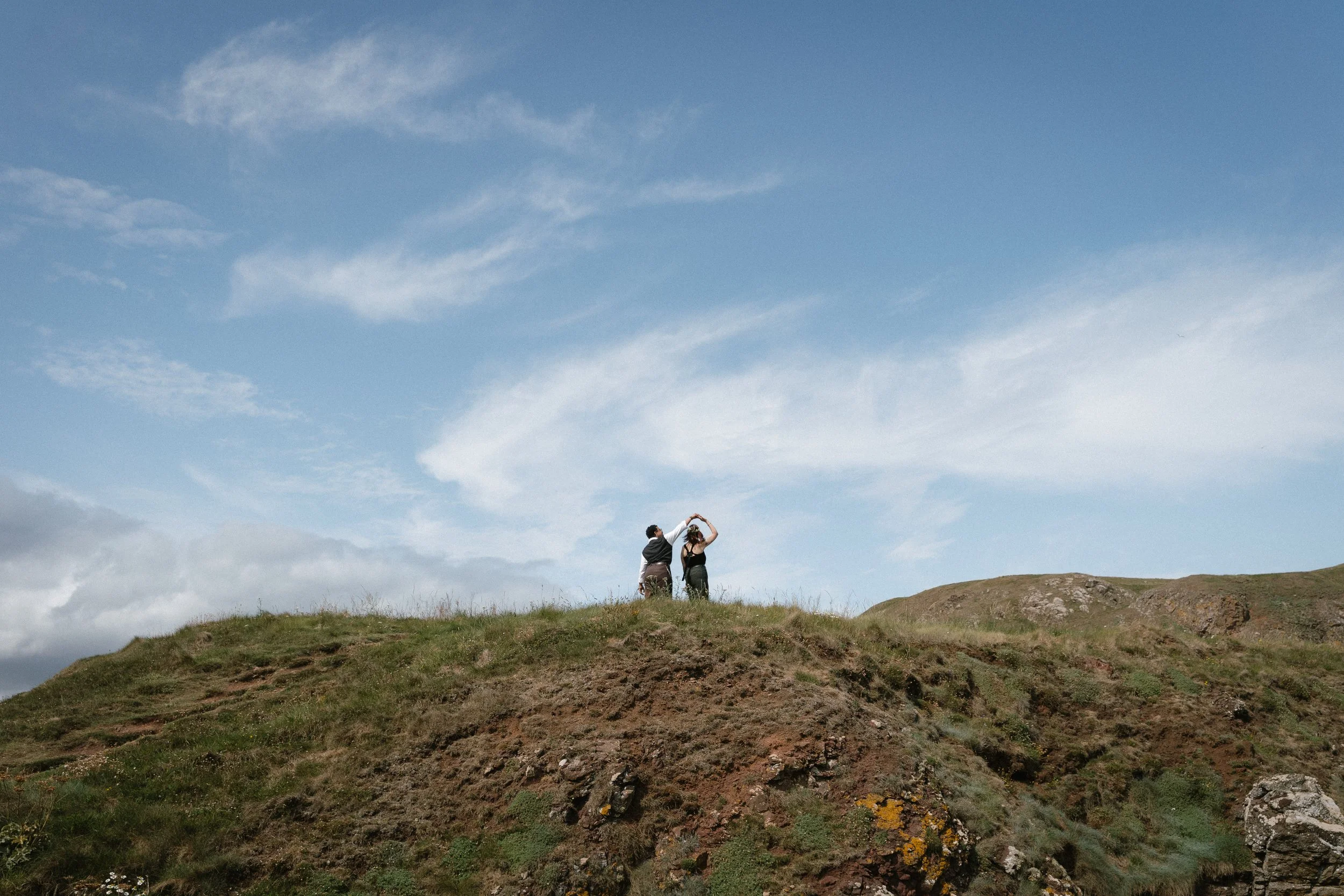 Two people standing on a grassy hilltop, holding hands above their heads against a blue sky with scattered clouds - captured by an Edinburgh wedding photographer