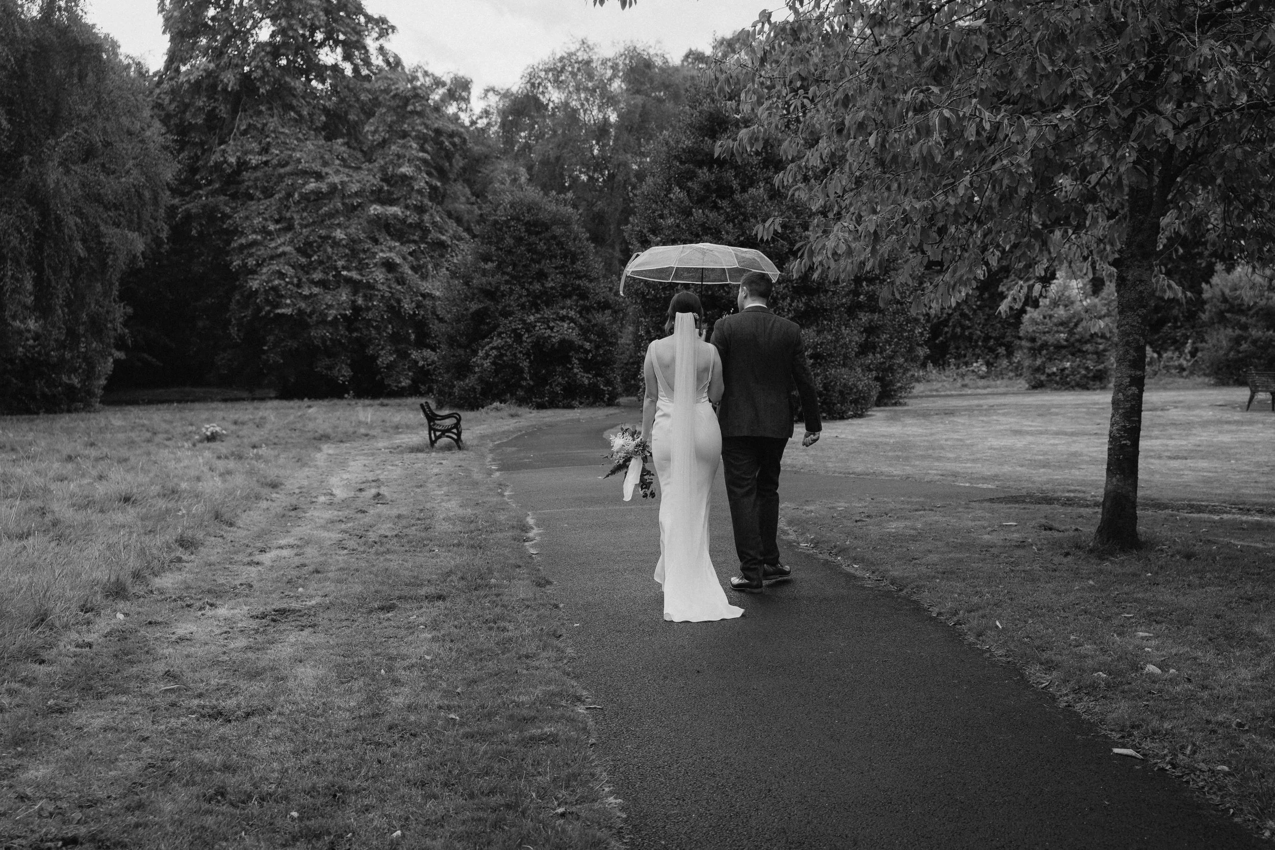 A bride and groom walk together under an umbrella on a path in a park, surrounded by trees and greenery, in black and white. Shot by an Edinburgh Wedding Photographer