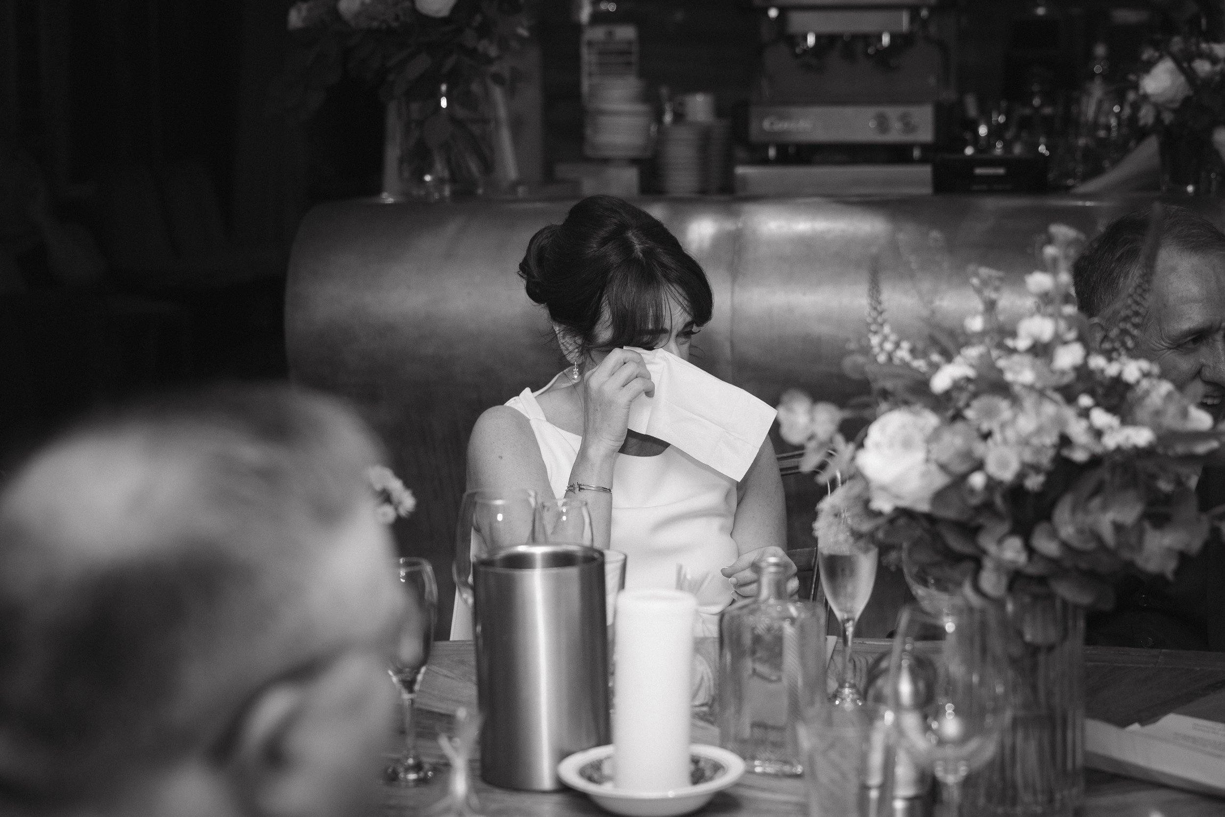 A woman sitting at a table, covering her face with a tissue, possibly crying or emotional, with other guests, glasses, and a flower arrangement around her. - captured by an Edinburgh wedding photographer