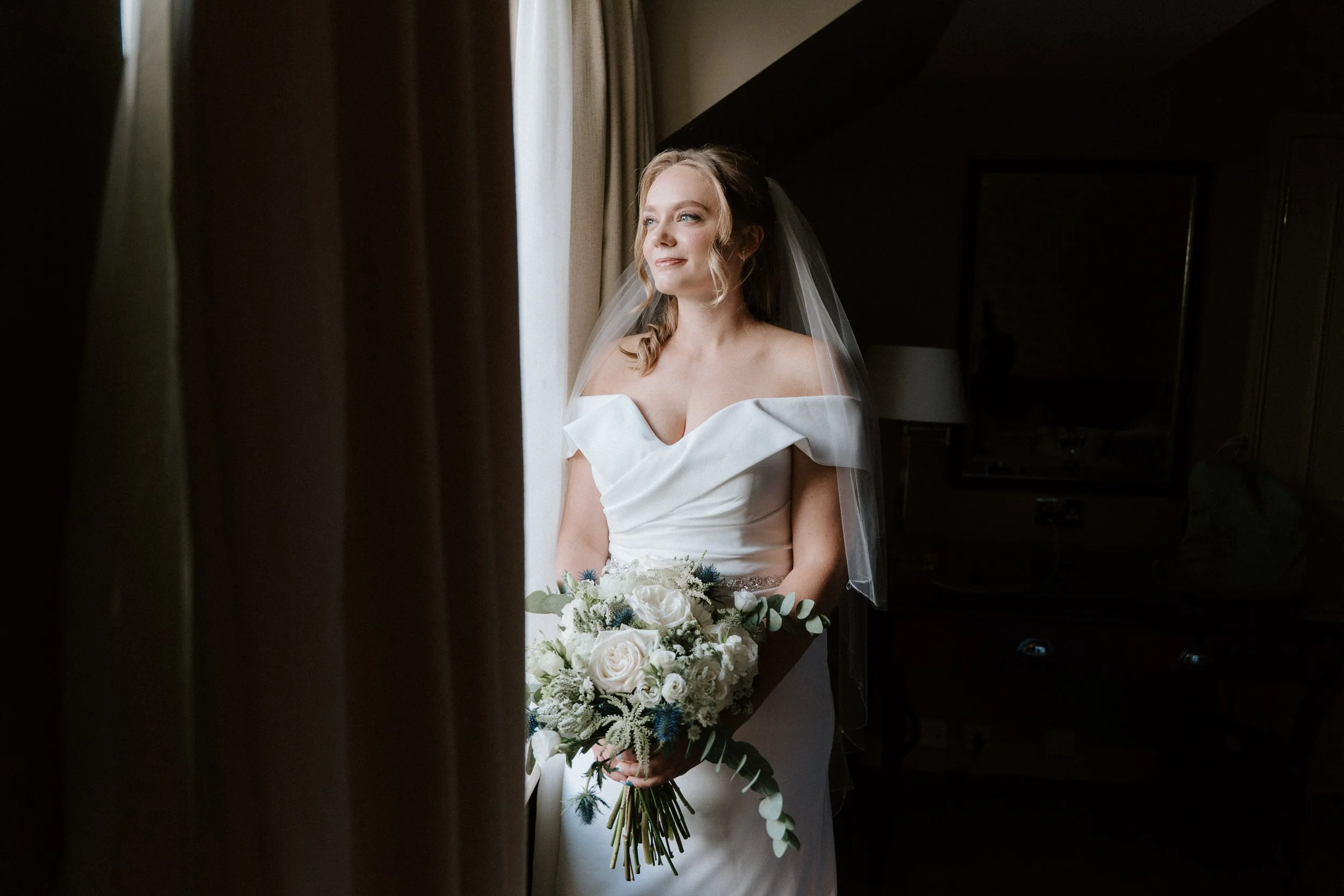 A bride in a white off-the-shoulder wedding dress holding a bouquet of white roses and greenery, standing near a window with curtains, looking out with a smile. - captured by an Edinburgh wedding photographer