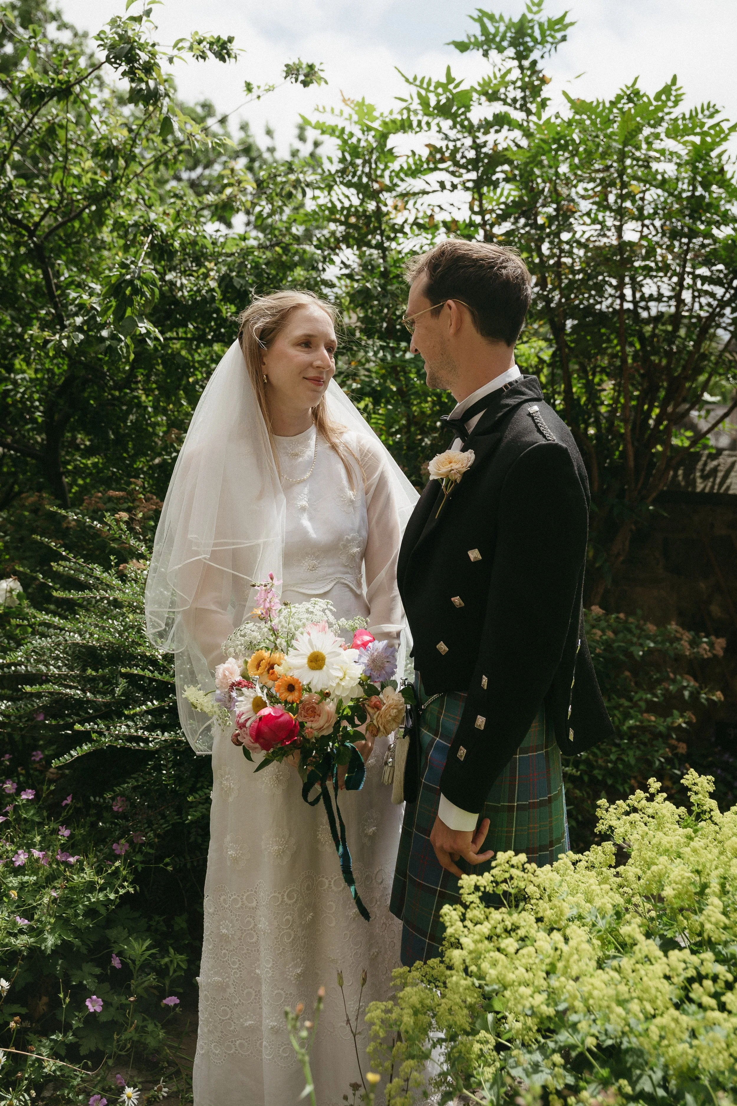 A bride and groom stand outdoors in a garden, facing each other during their wedding ceremony. The bride wears a white lace dress with a veil and holds a colorful bouquet. The groom wears a traditional Scottish kilt and a black jacket. They are surro