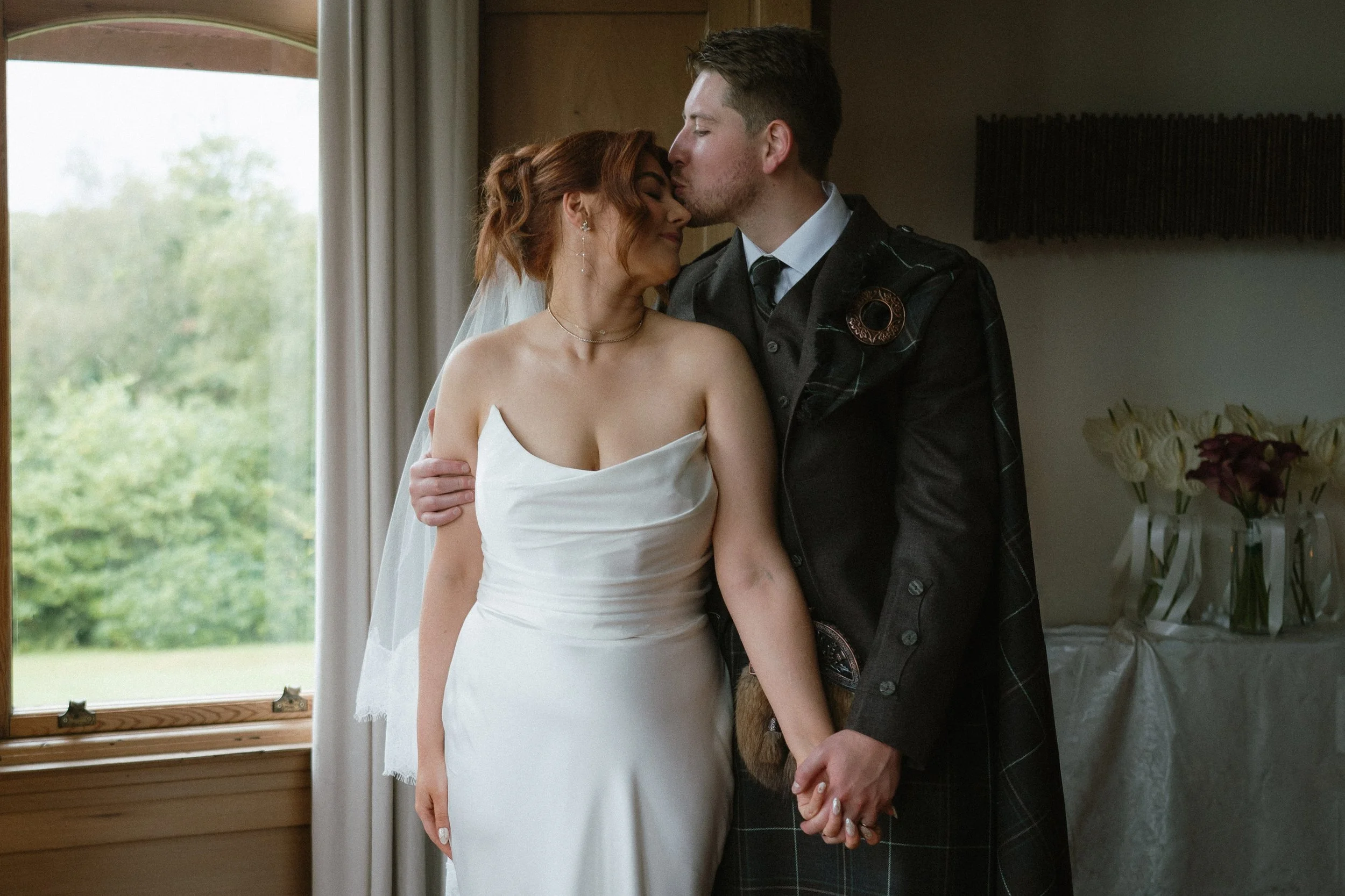 A bride and groom sharing a kiss in an indoor setting near a window with green scenery outside, holding hands with outdoors visible. - captured by an Edinburgh wedding photographer