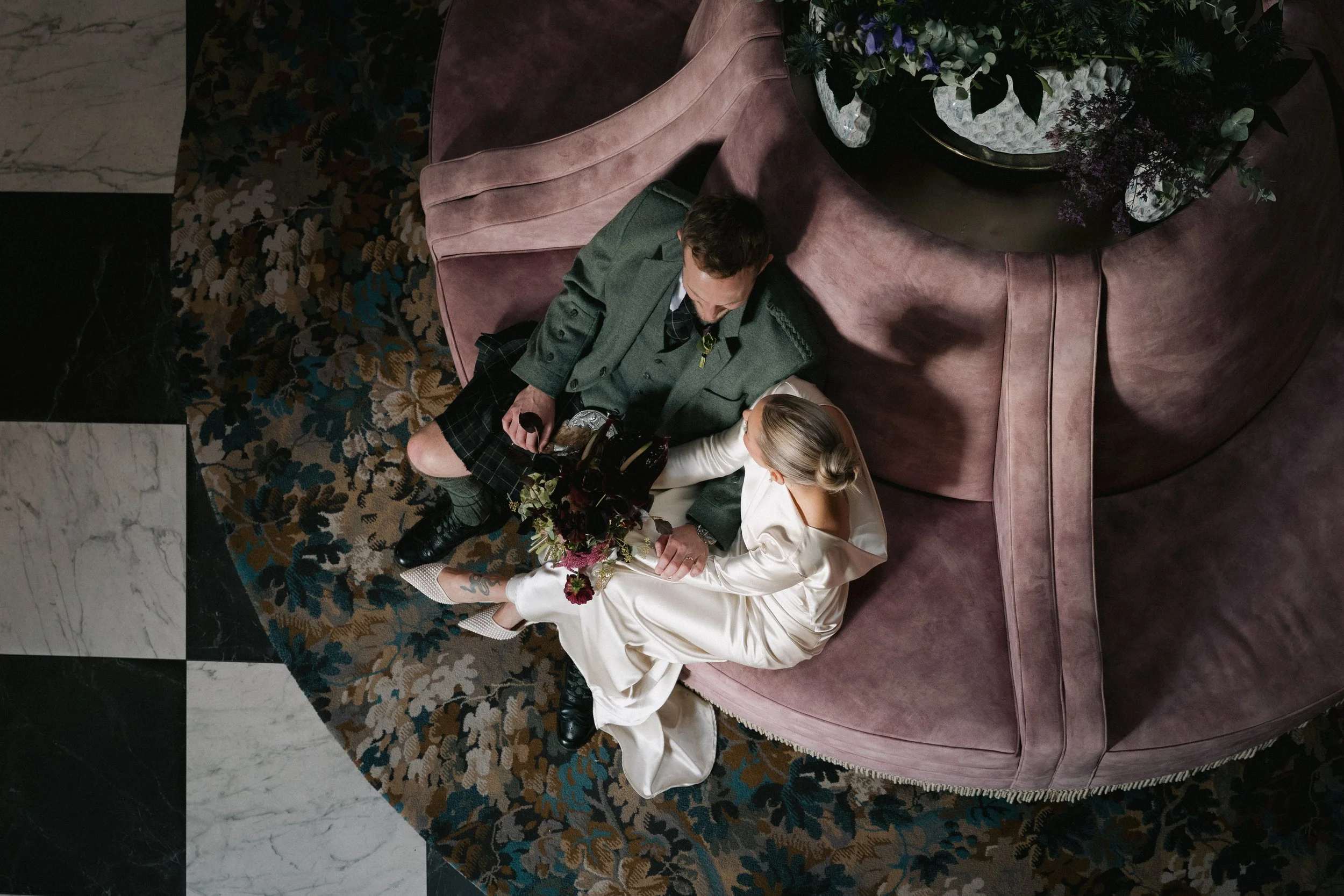 A couple sitting on a curved, pinkish velvet sofa, viewed from above. The woman is wearing a satin dress and holding a bouquet, while the man is dressed in a green blazer, patterned shirt, and shorts. They are on a floral patterned carpet with black 