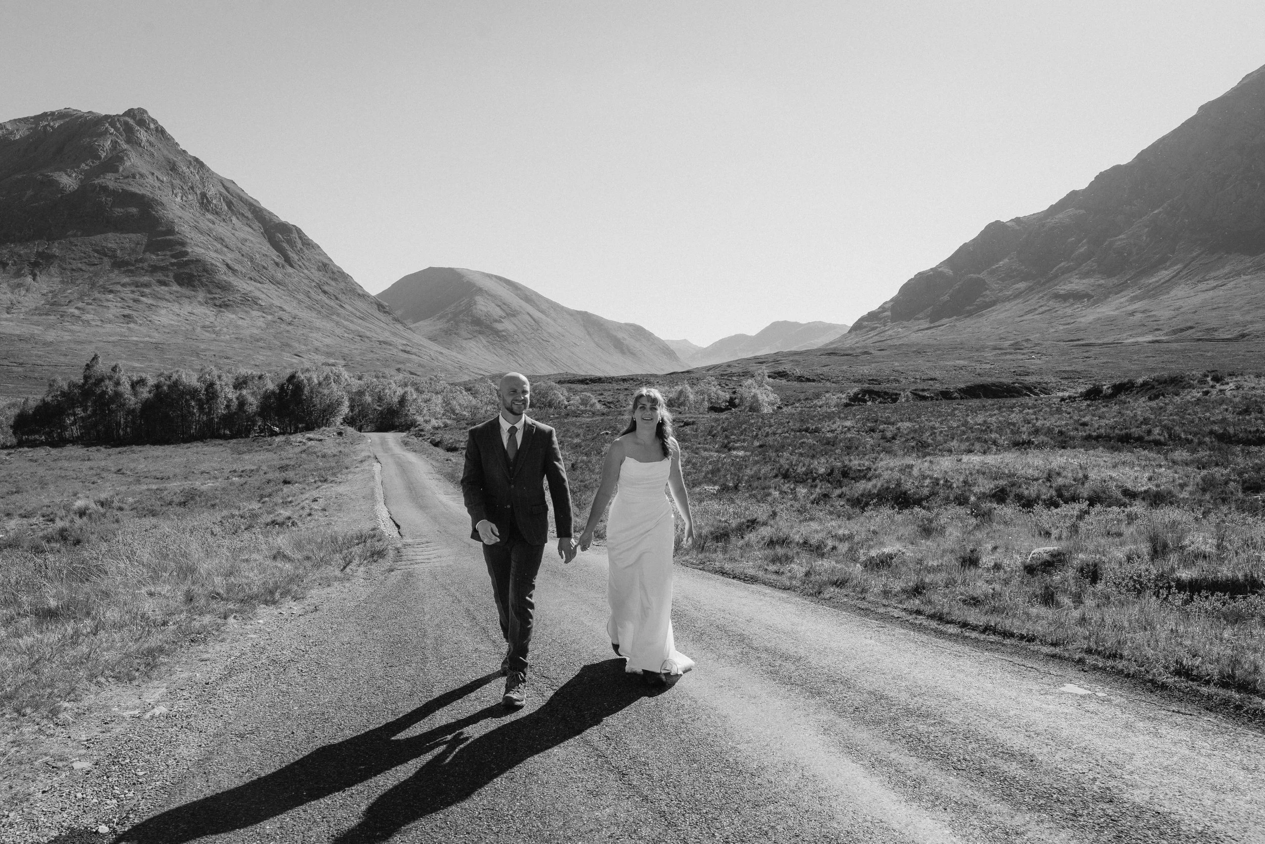 A black-and-white photo of a bride and groom walking hand-in-hand on a rural road surrounded by mountains and open land. - captured by an Edinburgh wedding photographer