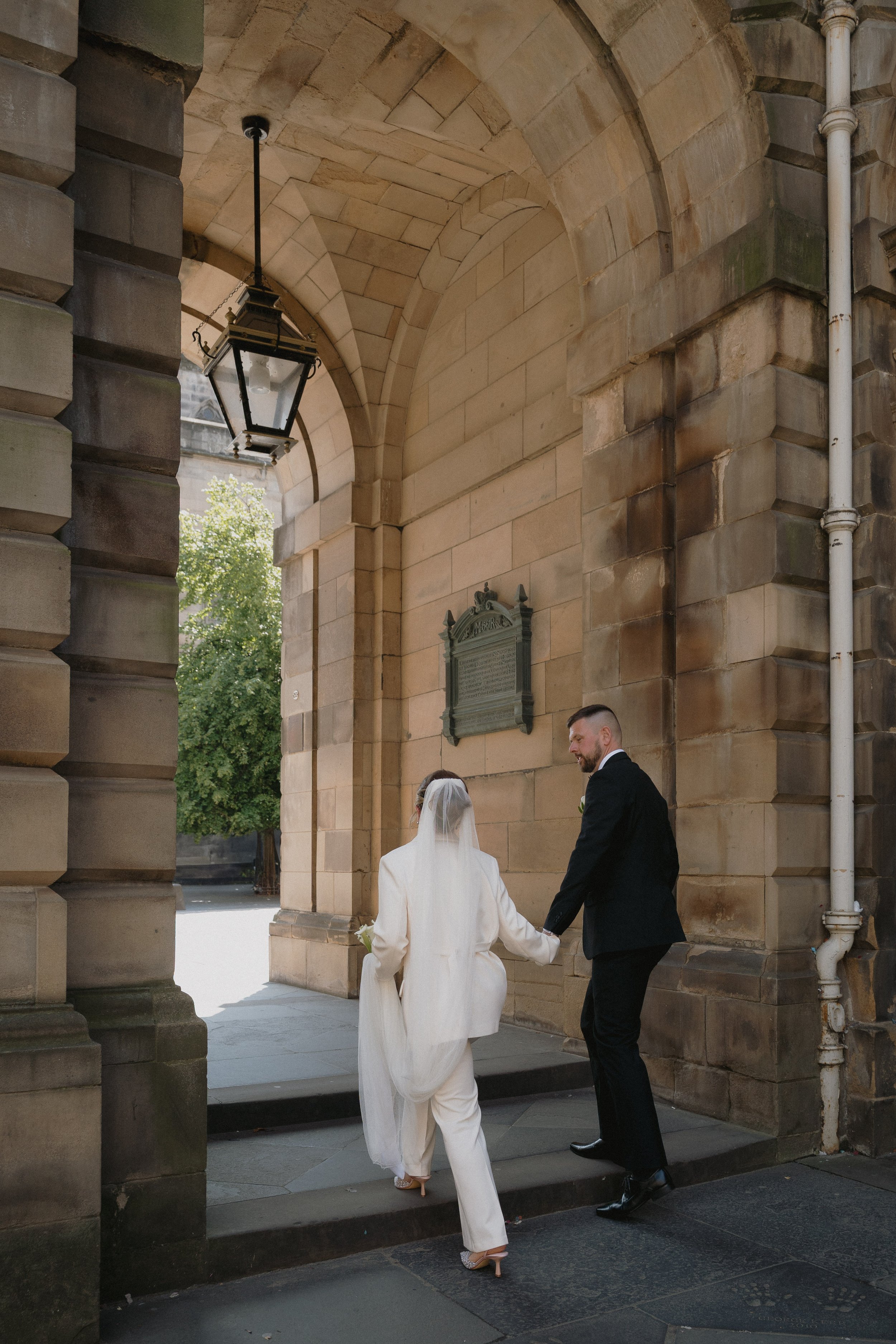 A bride and groom holding hands, walking through an archway of a historic stone building, with the bride dressed in a white suit and veil, and the groom in a black tuxedo. - captured by an Edinburgh wedding photographer