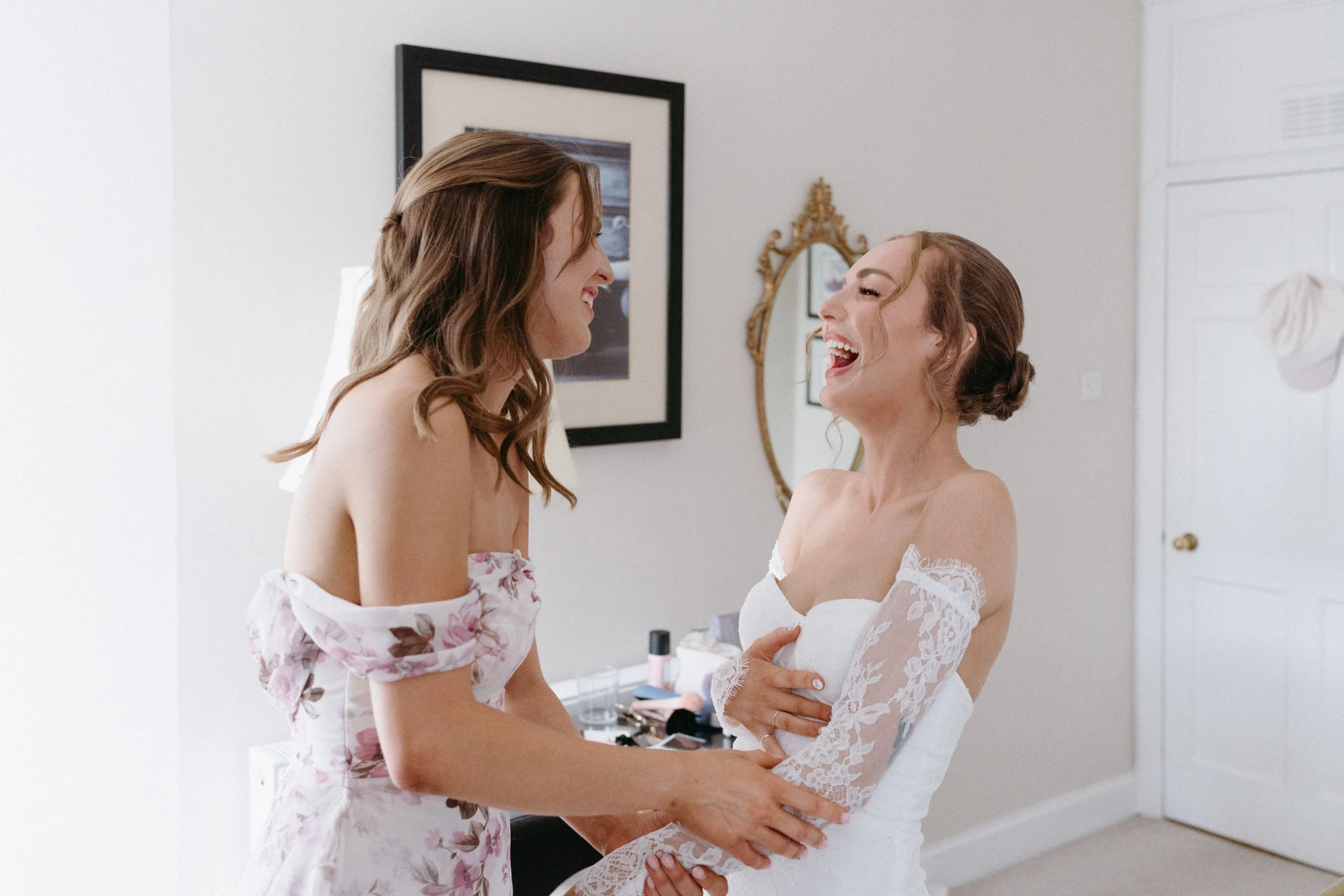 Two women, one in a floral dress and one in a white wedding gown, laughing and holding hands in a room with a mirror and framed pictures on the wall - captured by an Edinburgh wedding photographer