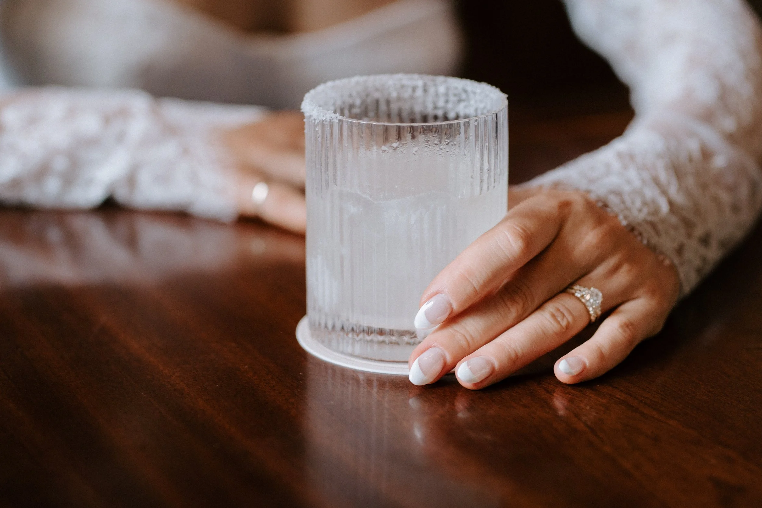 A woman with a wedding ring on her finger resting her hand on a wooden table, near a textured glass of water with frost on the rim, wearing a lace sleeve. - captured by an Edinburgh wedding photographer