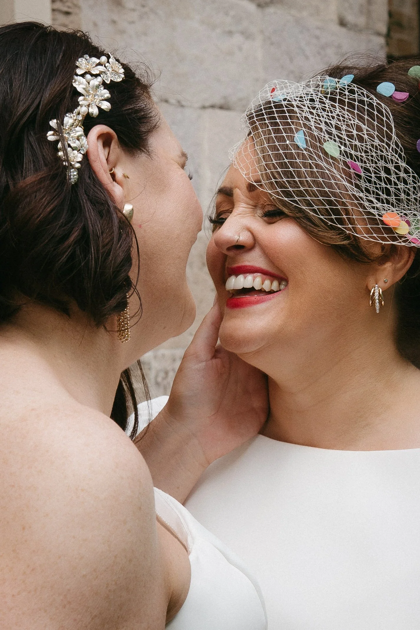 Two women celebrating at a wedding, smiling and close face-to-face, wearing white dresses, with one wearing a floral hairpiece and the other a birdcage veil with colorful confetti. - captured by an Edinburgh wedding photographer