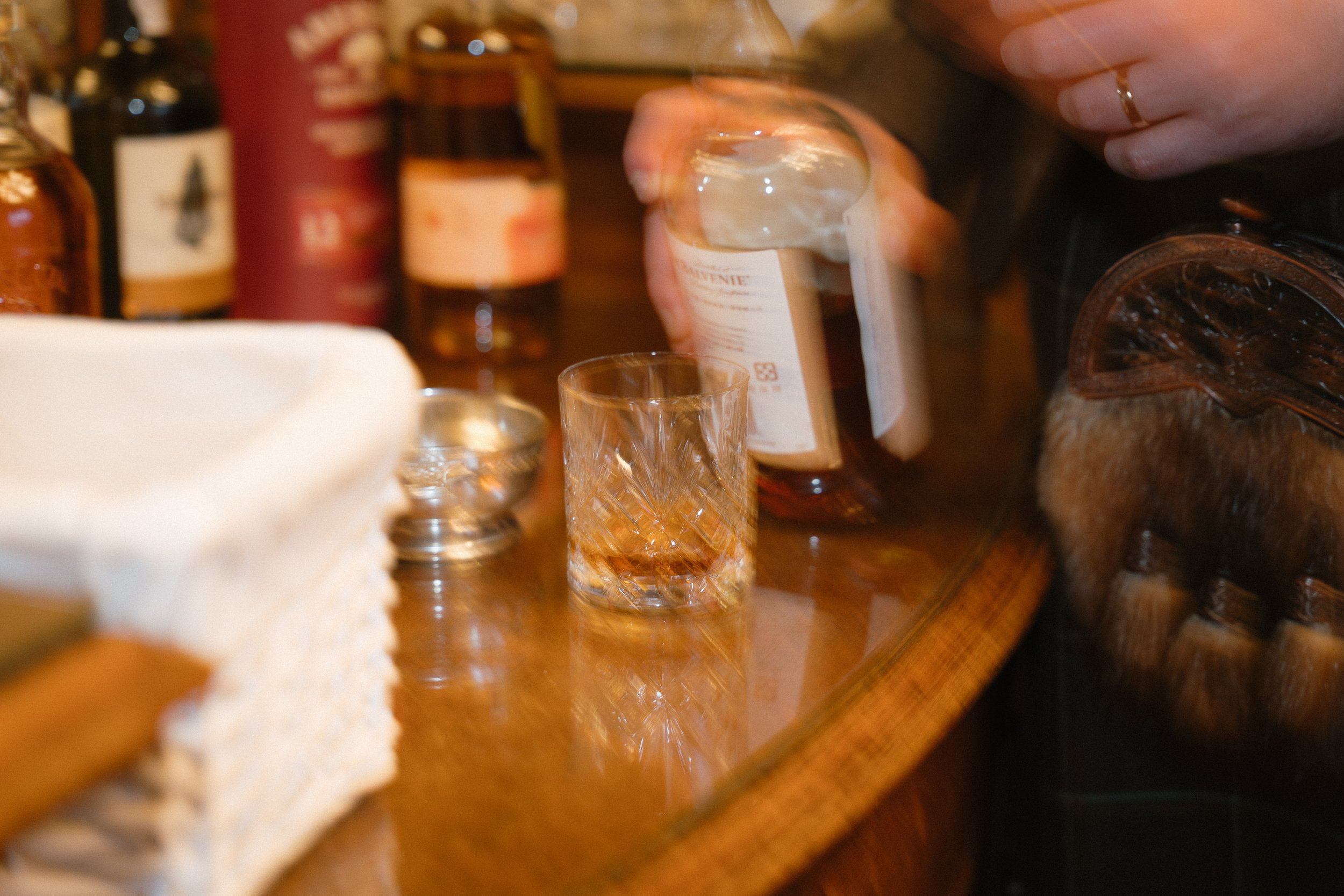 Close-up of a person pouring whisky into a glass on a bar counter with bottles of alcohol and a white cloth in the background. - captured by an Edinburgh wedding photographer