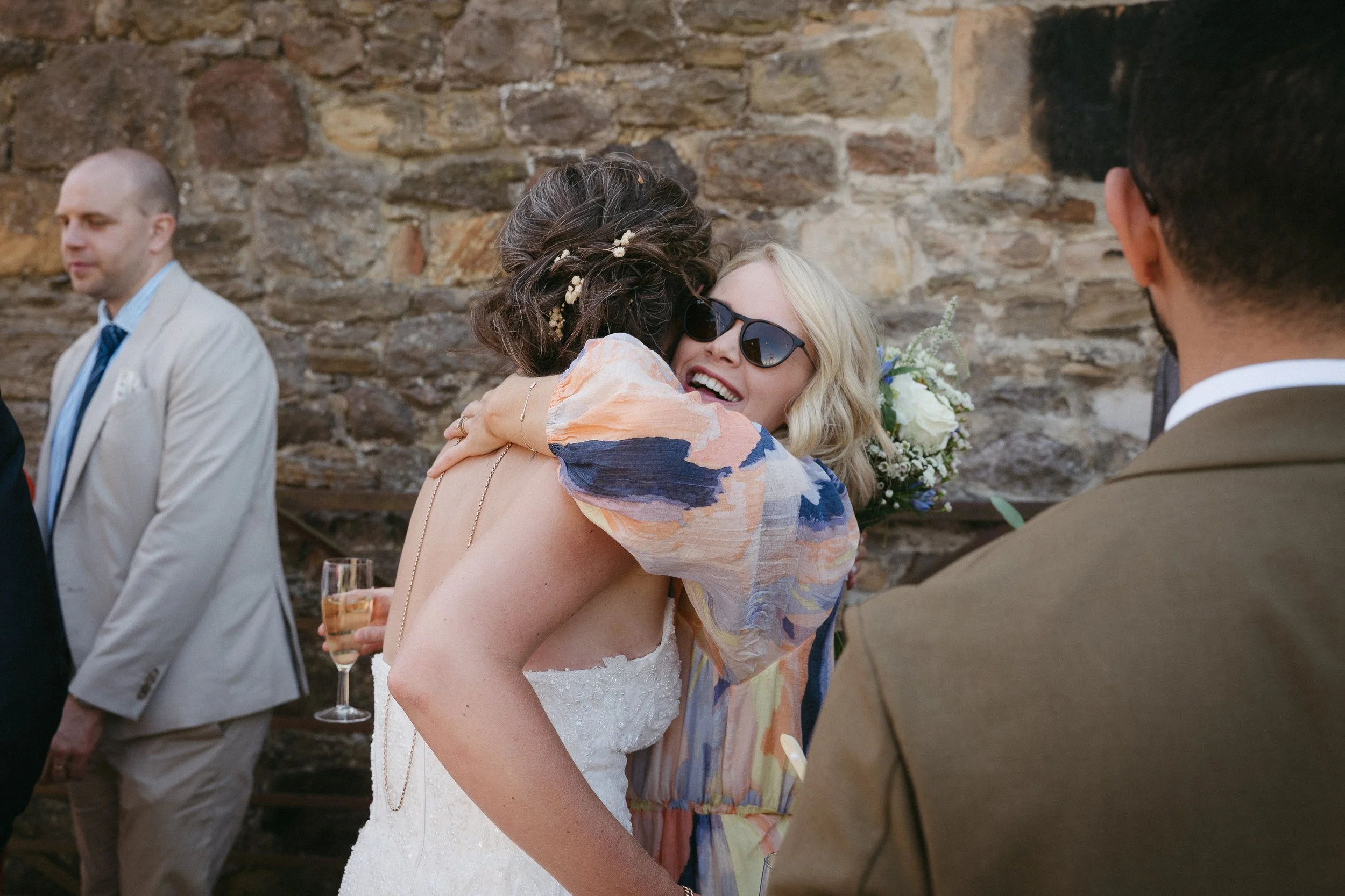 A woman in a wedding dress, wearing sunglasses, embracing another woman with blonde hair and sunglasses at a wedding reception outdoors, with a stone wall in the background. - captured by an Edinburgh wedding photographer