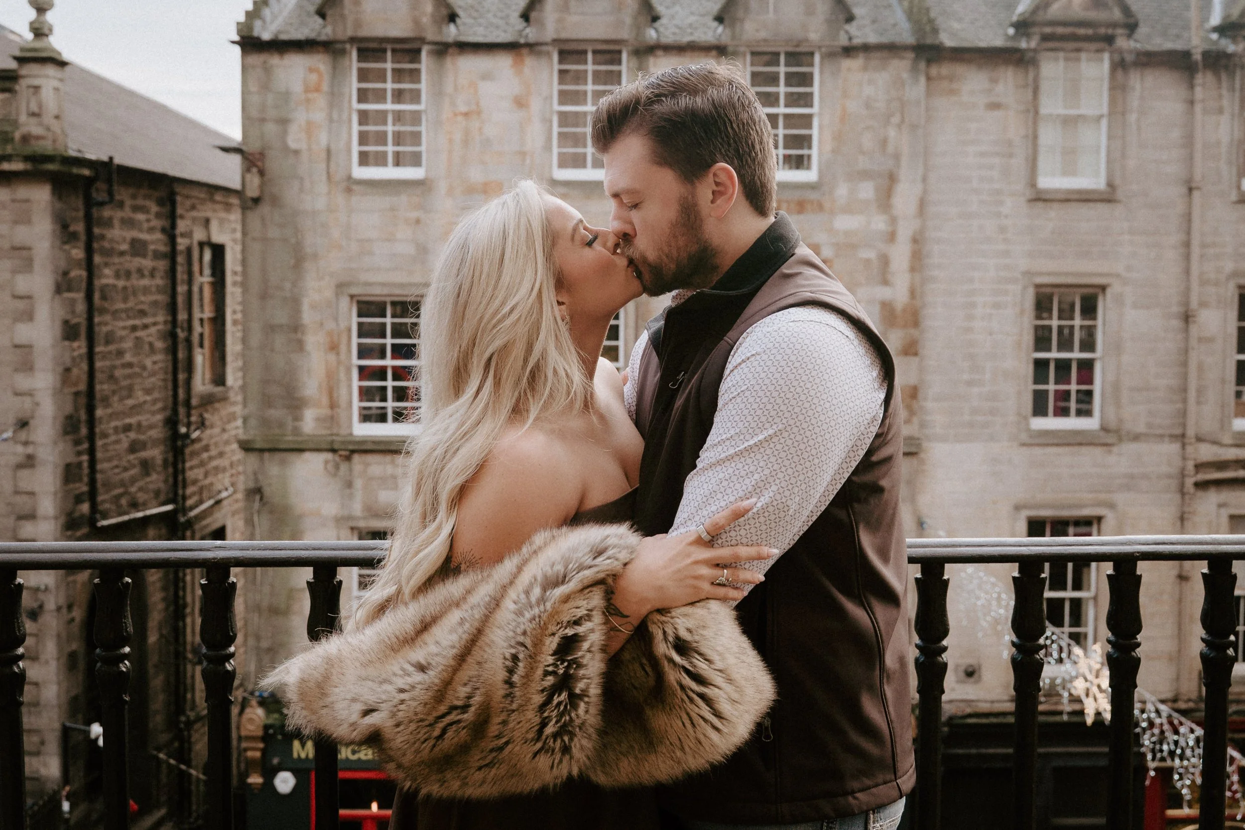 A couple sharing a kiss on a balcony, with an old stone building in the background, the woman wearing a fur coat and the man in a vest and long-sleeve shirt. - captured by an Edinburgh wedding photographer
