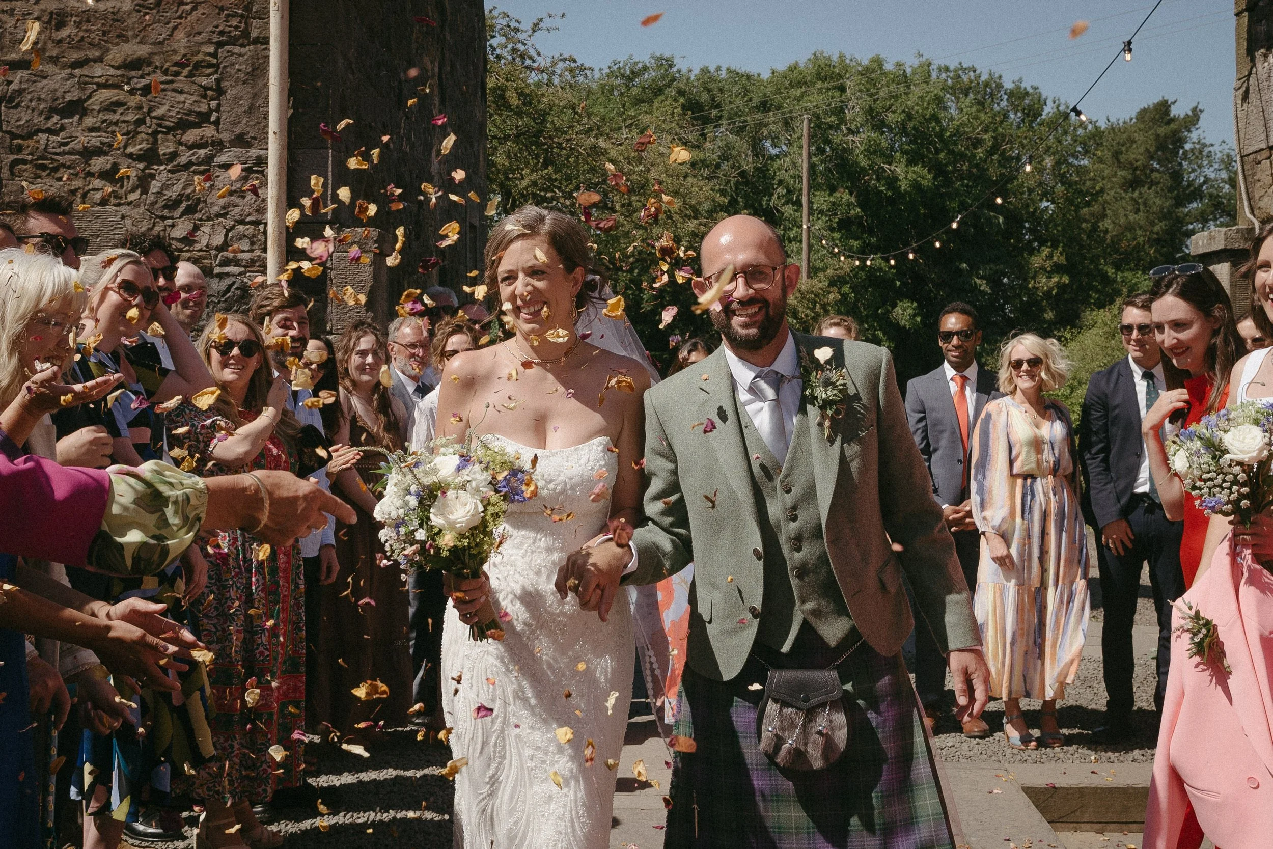 Bride and groom walking through a crowd throwing flower petals, outdoor wedding celebration on sunny day. - captured by an Edinburgh wedding photographer