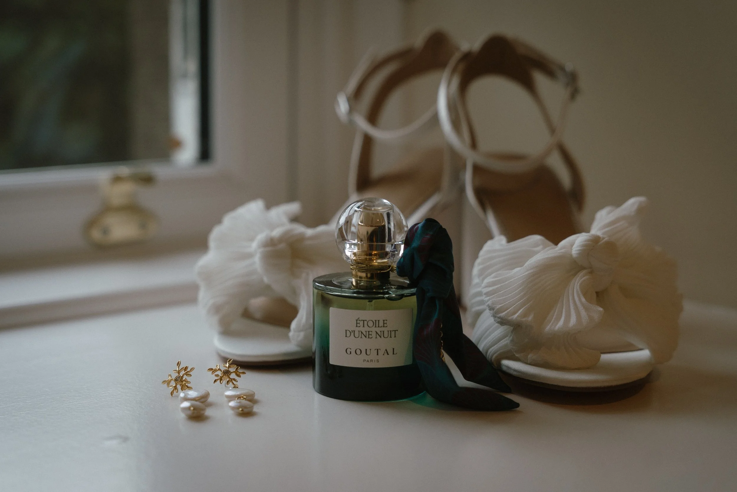A bottle of Goutal Paris perfume labeled 'Étoile D'une Nuit' on a white surface, with white sandals, gold earrings, and a black scarf nearby, in front of a window. - captured by an Edinburgh wedding photographer