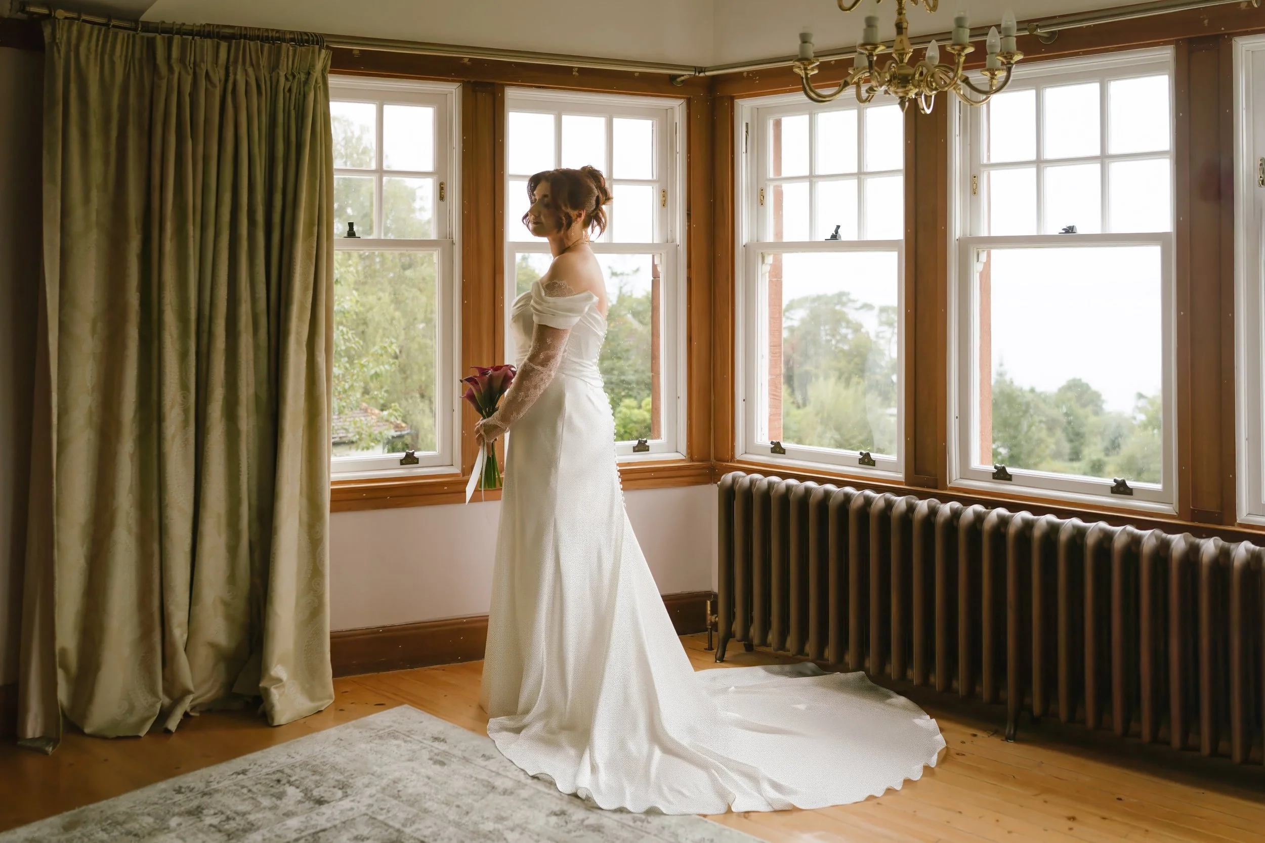 A bride in a white wedding gown holding a bouquet, standing in front of large windows with wooden frames inside a room with wooden floors and a radiator. - captured by an Edinburgh wedding photographer