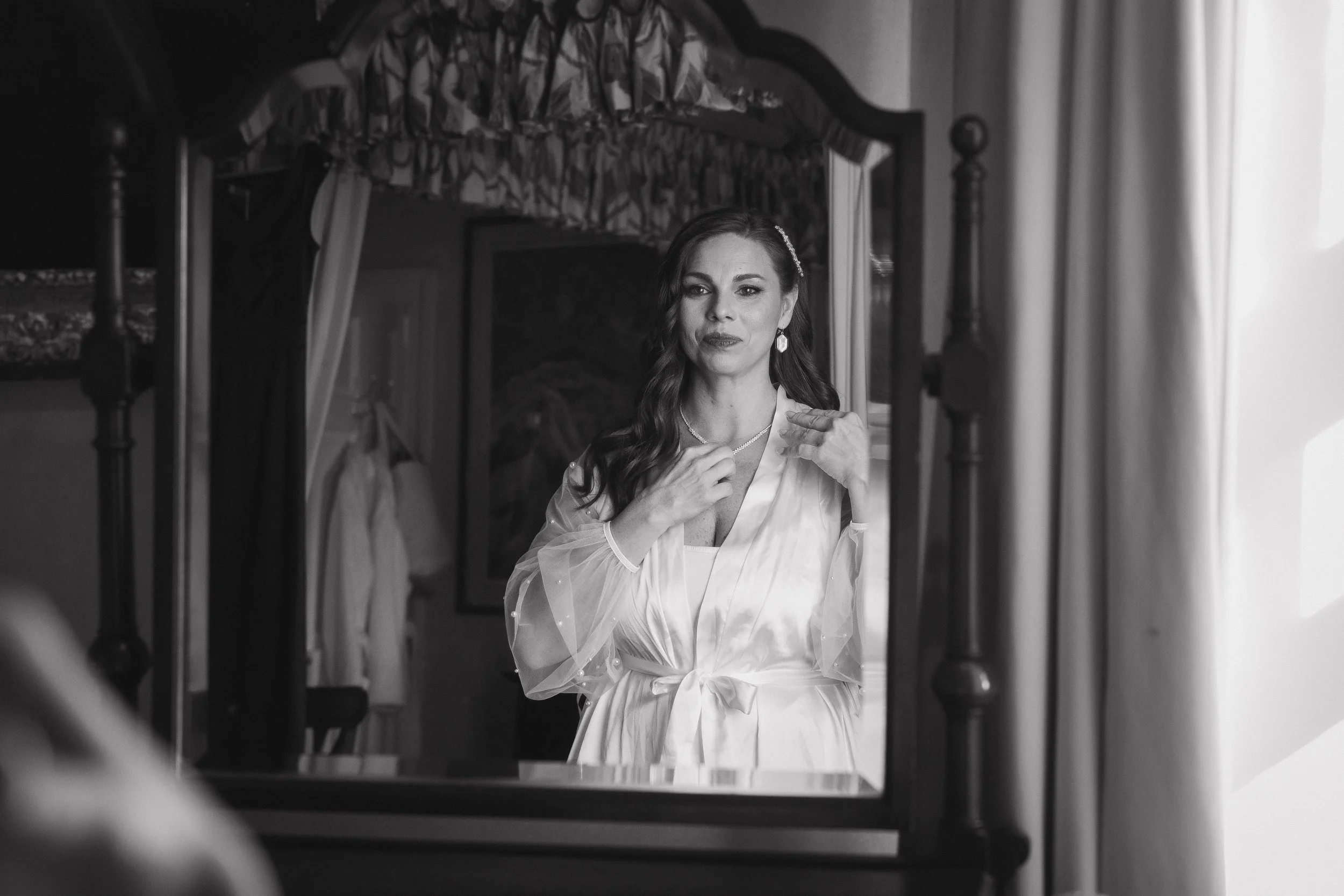 A woman with long dark hair and earrings looking at her reflection in a mirror, adjusting a necklace, in a room with a bed and curtains. - captured by an Edinburgh wedding photographer