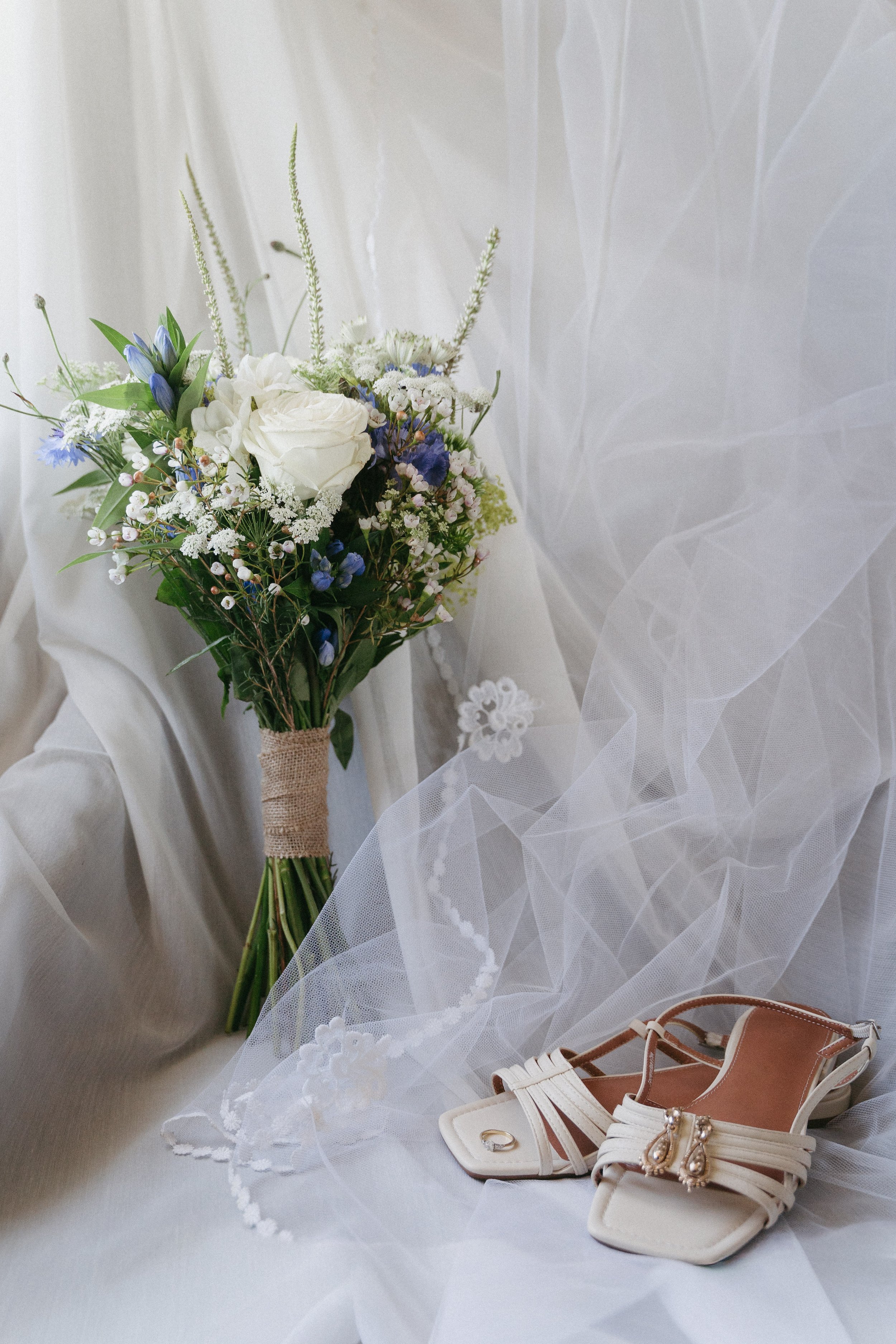 A bouquet of white and purple flowers, a pair of white shoes with jewelry, and a sheer white veil on a light-colored fabric surface. - captured by an Edinburgh wedding photographer