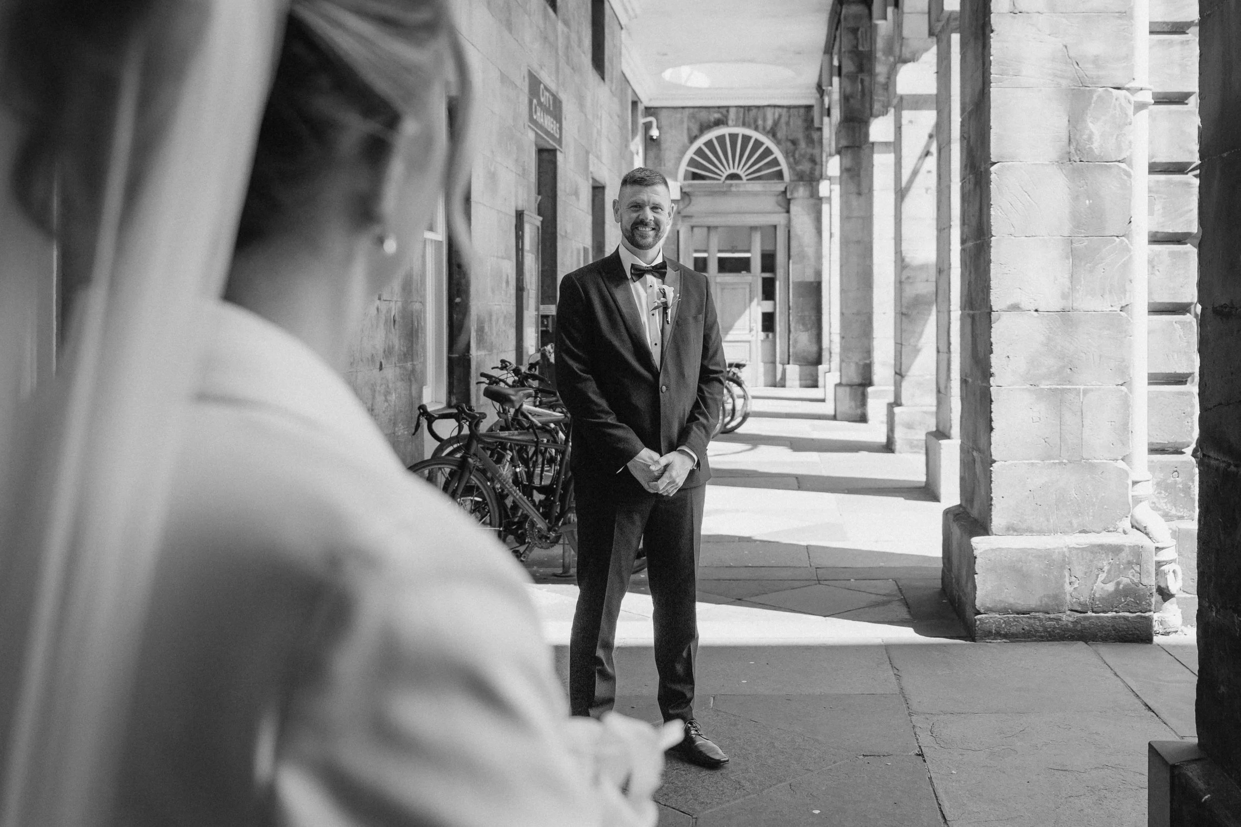 A groom in a tuxedo stands under a stone archway, smiling at the bride, who is partially visible in the foreground. The scene is in black and white, with bicycles parked along the building wall behind him.