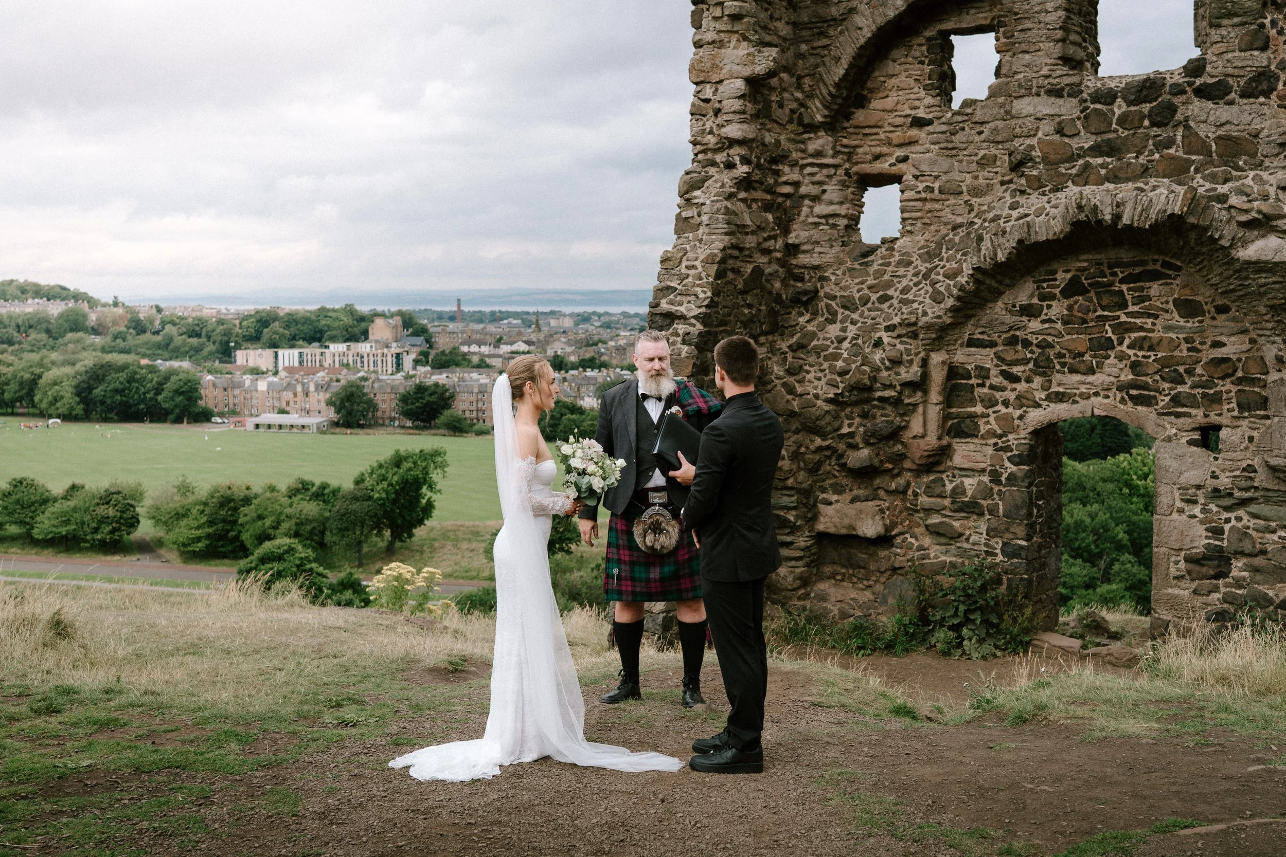 A couple gets married outdoors with a scenic view of a city and green landscape, officiated by a man in a kilt and traditional attire. The bride is in a white wedding dress holding a bouquet, and the groom is in a black suit. - captured by an Edinbur