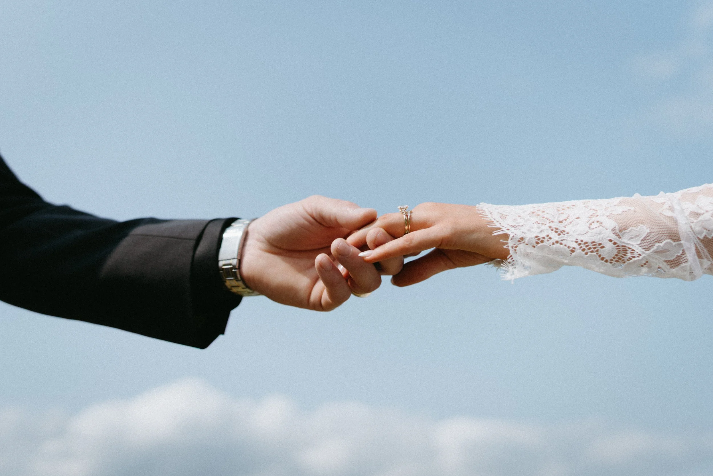 Close-up of a couple holding hands during a wedding, with one person wearing a black suit and the other in a lace wedding dress, against a blue sky background. - captured by an Edinburgh wedding photographer