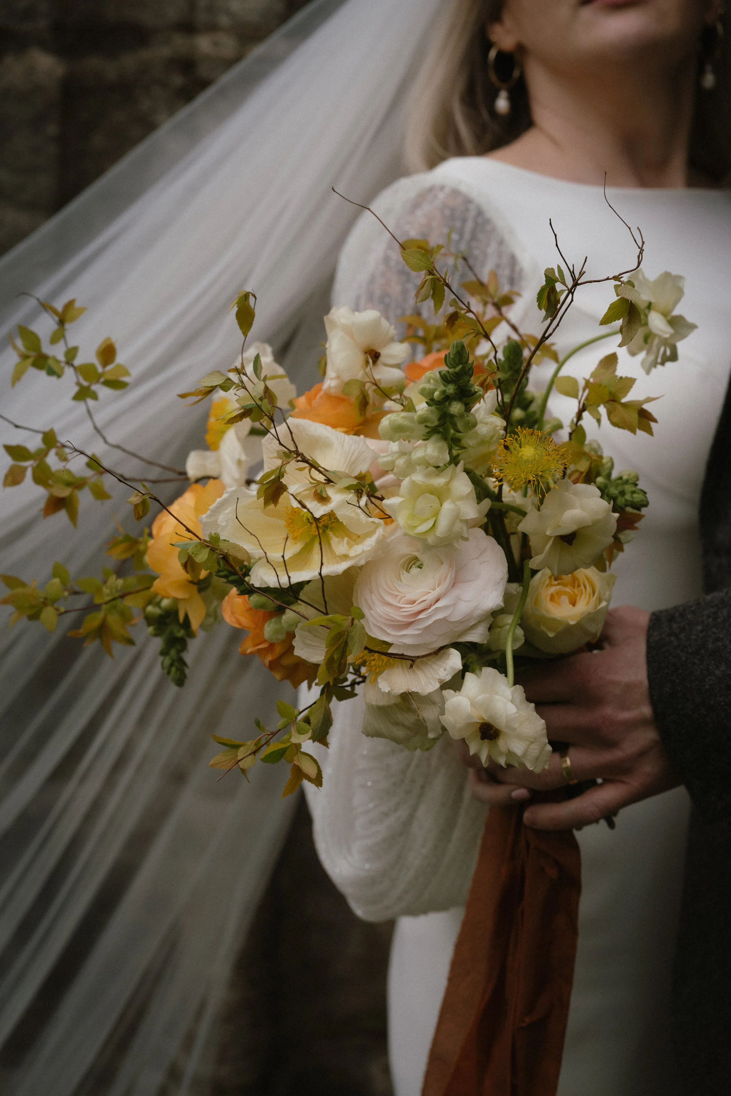 Bride wearing a white dress and veil holding a bouquet of peach, cream, and green flowers with greenery, during a wedding ceremony. - captured by an Edinburgh wedding photographer