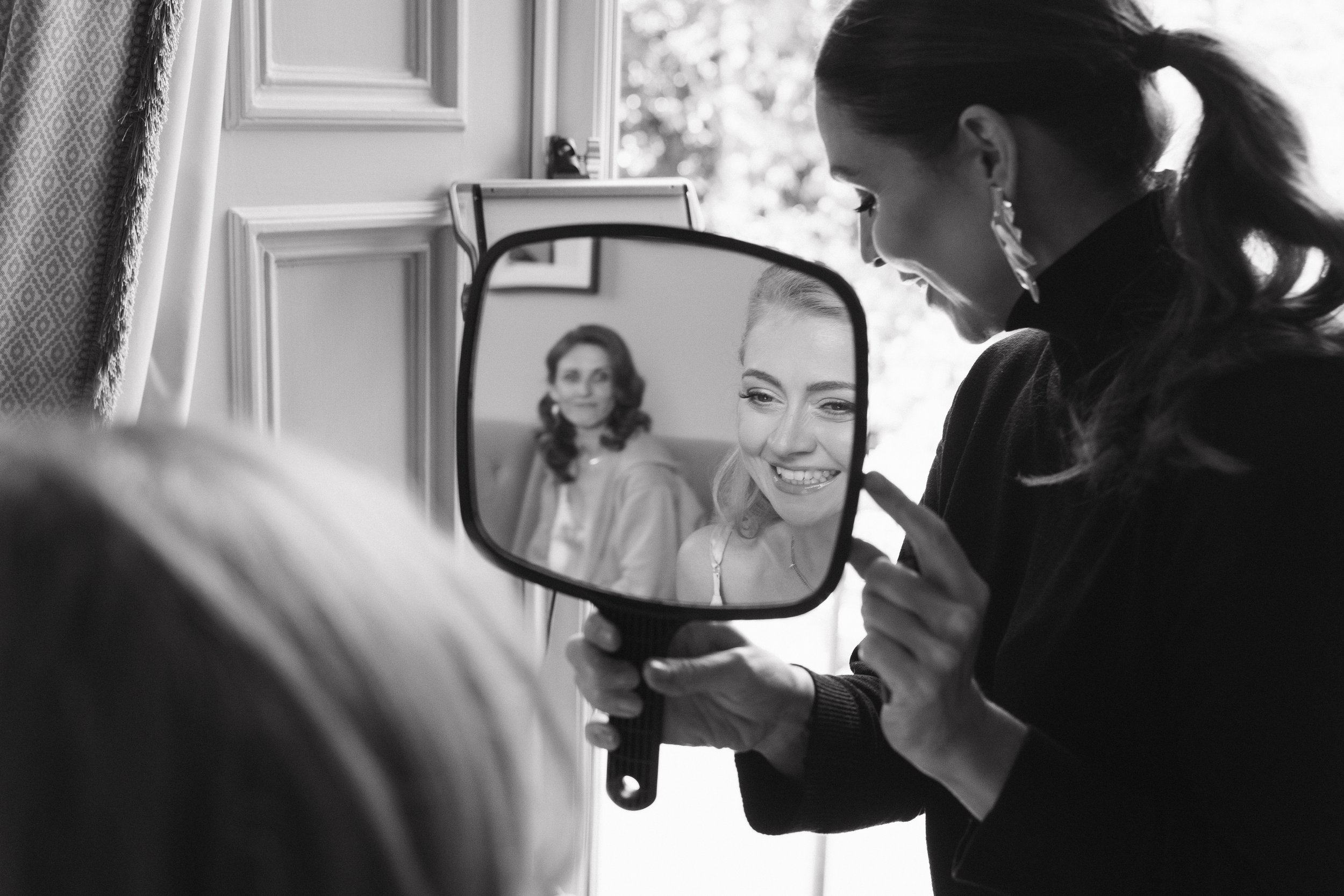 A woman is holding a mirror showing her reflection as she gets ready, with another woman in the background looking on, in a room with a window and patterned curtain. - captured by an Edinburgh wedding photographer
