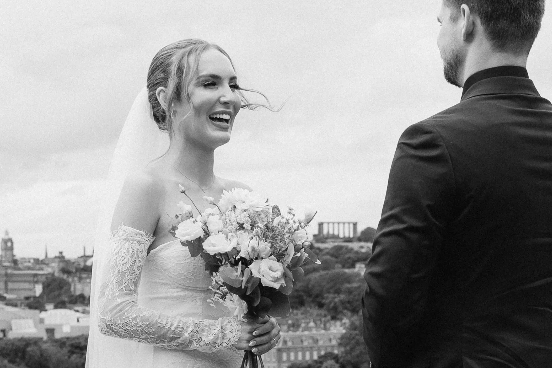 Black and white photo of a bride and groom during a wedding ceremony outdoors. The bride is smiling and holding a bouquet of flowers, while the groom is facing her, partially visible from behind. - captured by an Edinburgh wedding photographer