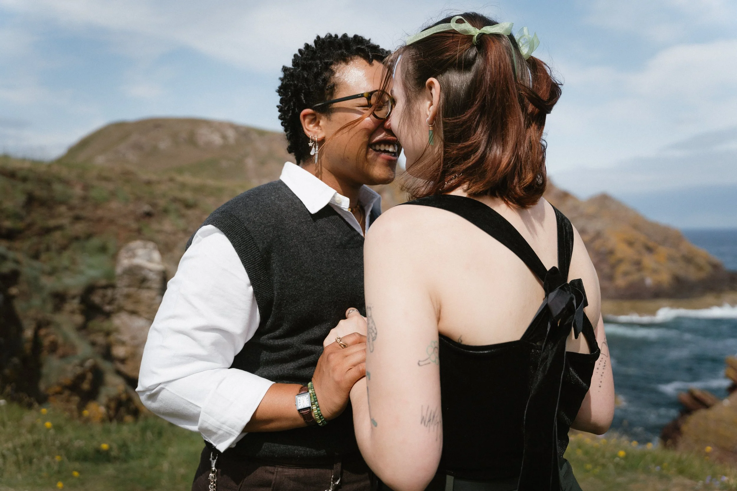 Two women face each other close, smiling, outdoors near the coast with rocky hills and ocean in the background - captured by an Edinburgh wedding photographer