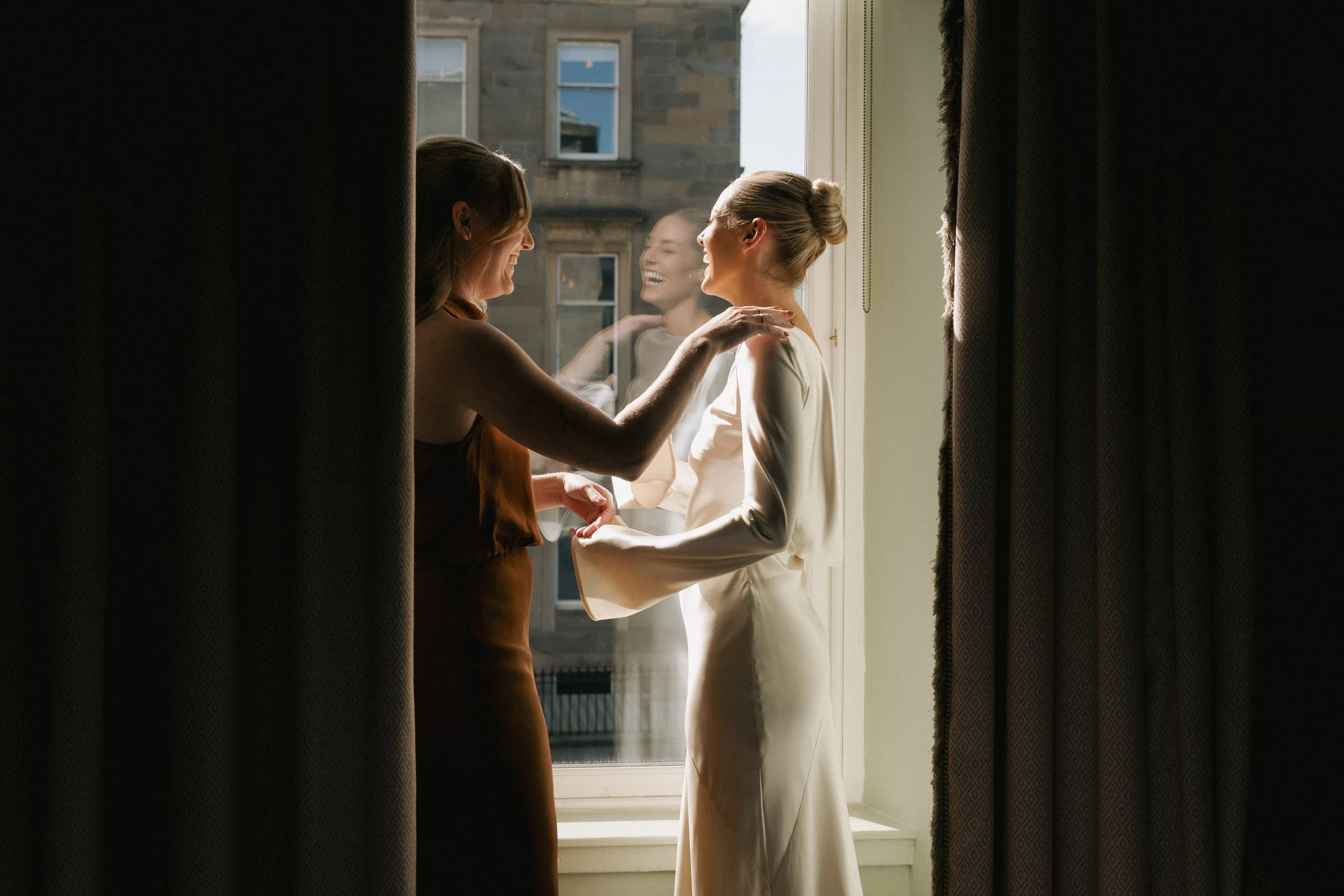 Two women, one in a white wedding dress and the other in a brown dress, share a joyful moment by a window, with their reflection visible on the glass. - captured by an Edinburgh wedding photographer