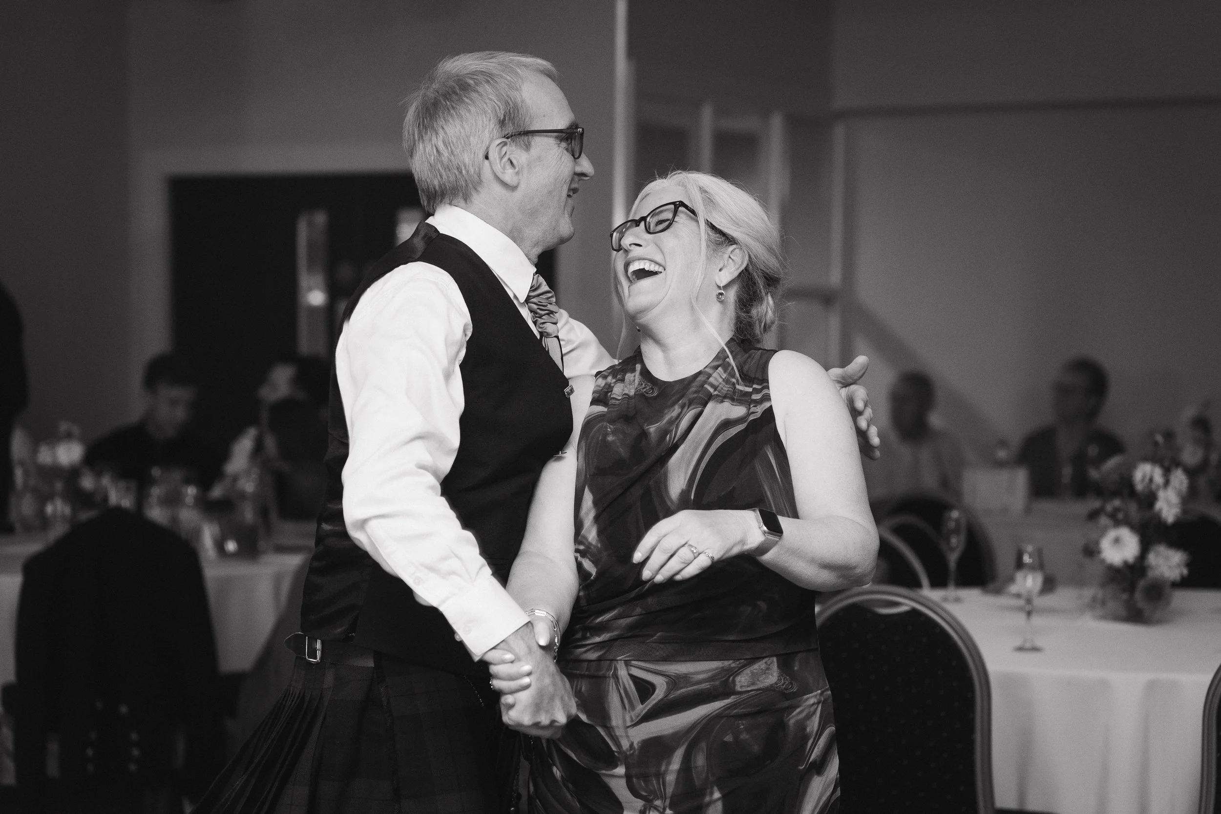 A black and white photo of a man in a vest and tie dancing with a woman in a sleeveless dress, both wearing glasses, smiling and laughing in a banquet hall. - captured by an Edinburgh wedding photographer