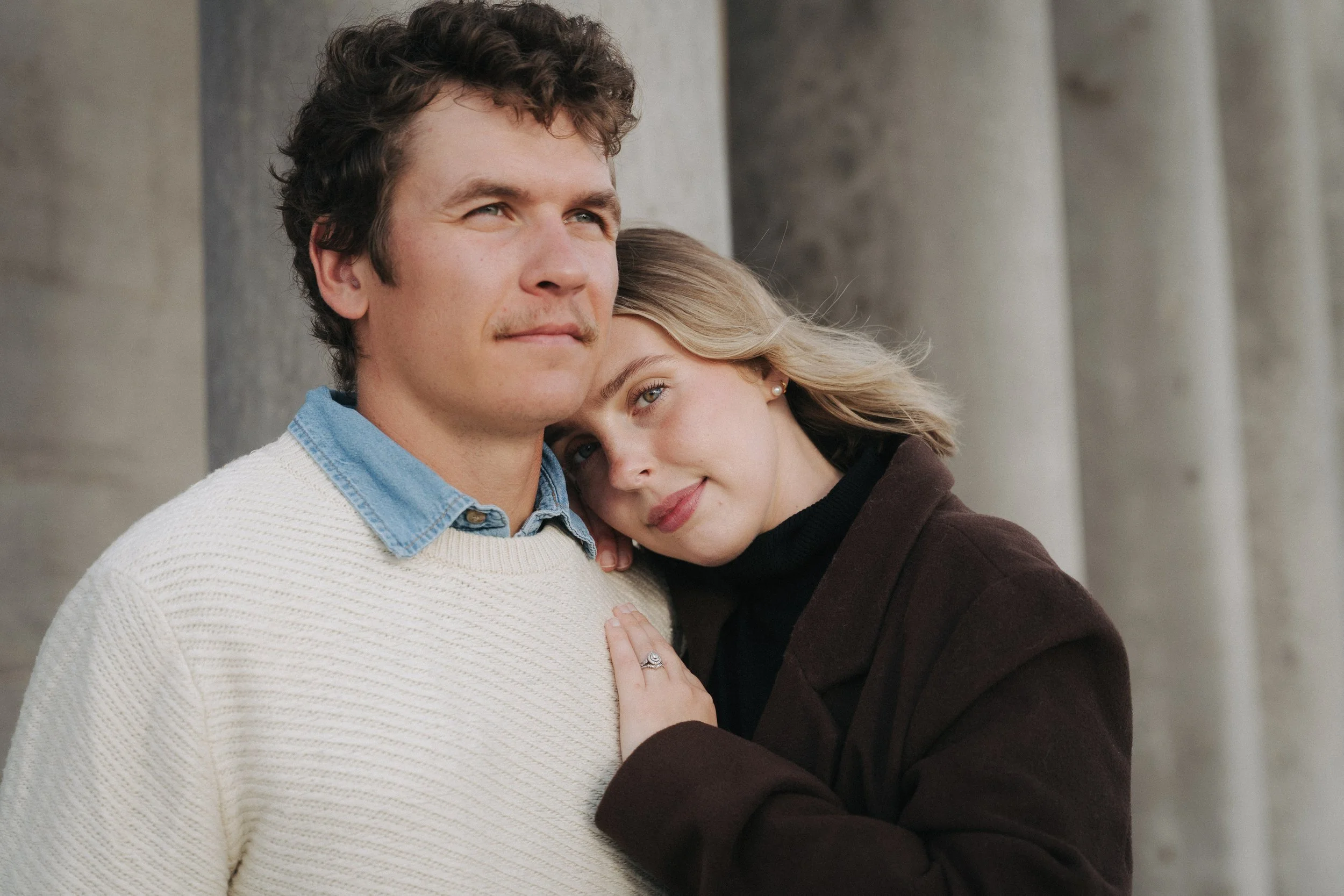A young man and woman embrace each other, the woman resting her head on the man's shoulder and smiling softly, with a background of large stone columns.
