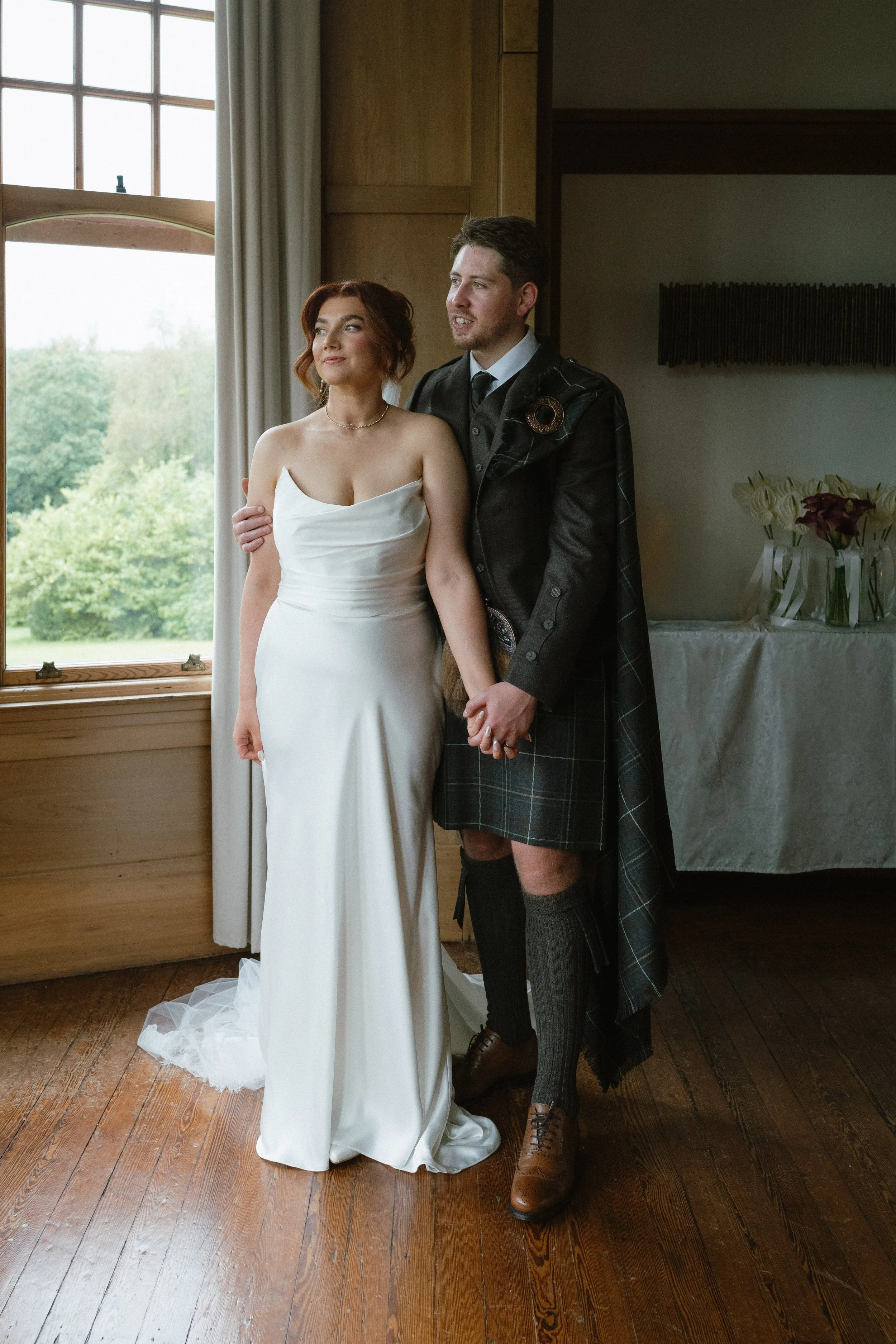 A bride in a white wedding gown and a groom in traditional Scottish attire hold hands near a window, standing on a wooden floor inside a room. - captured by an Edinburgh wedding photographer