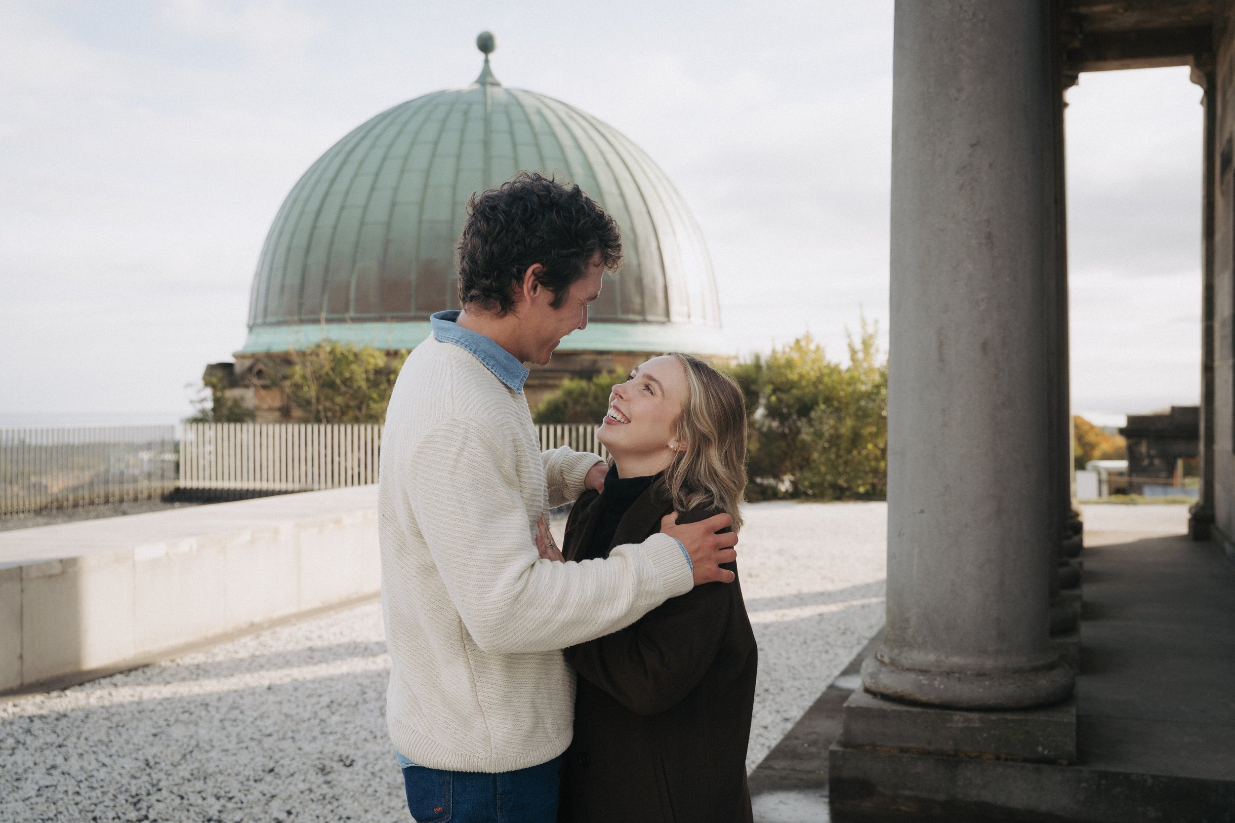 A couple smiling and embracing each other outdoors near a building with classical columns, with a dome structure in the background. - captured by an Edinburgh wedding photographer