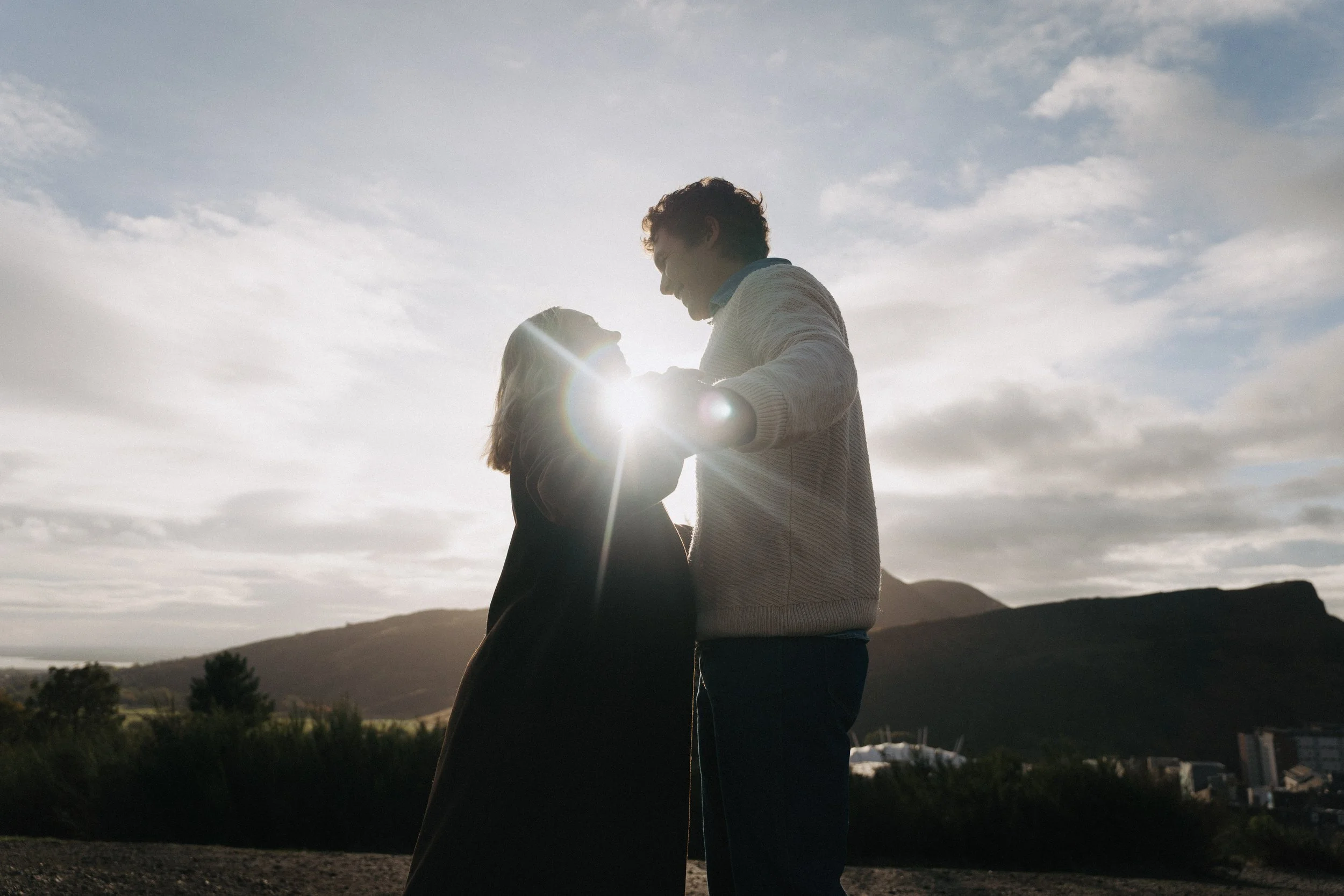 A couple standing close together outdoors during sunset, holding hands and smiling at each other with mountains and cloudy sky in the background. - captured by an Edinburgh wedding photographer
