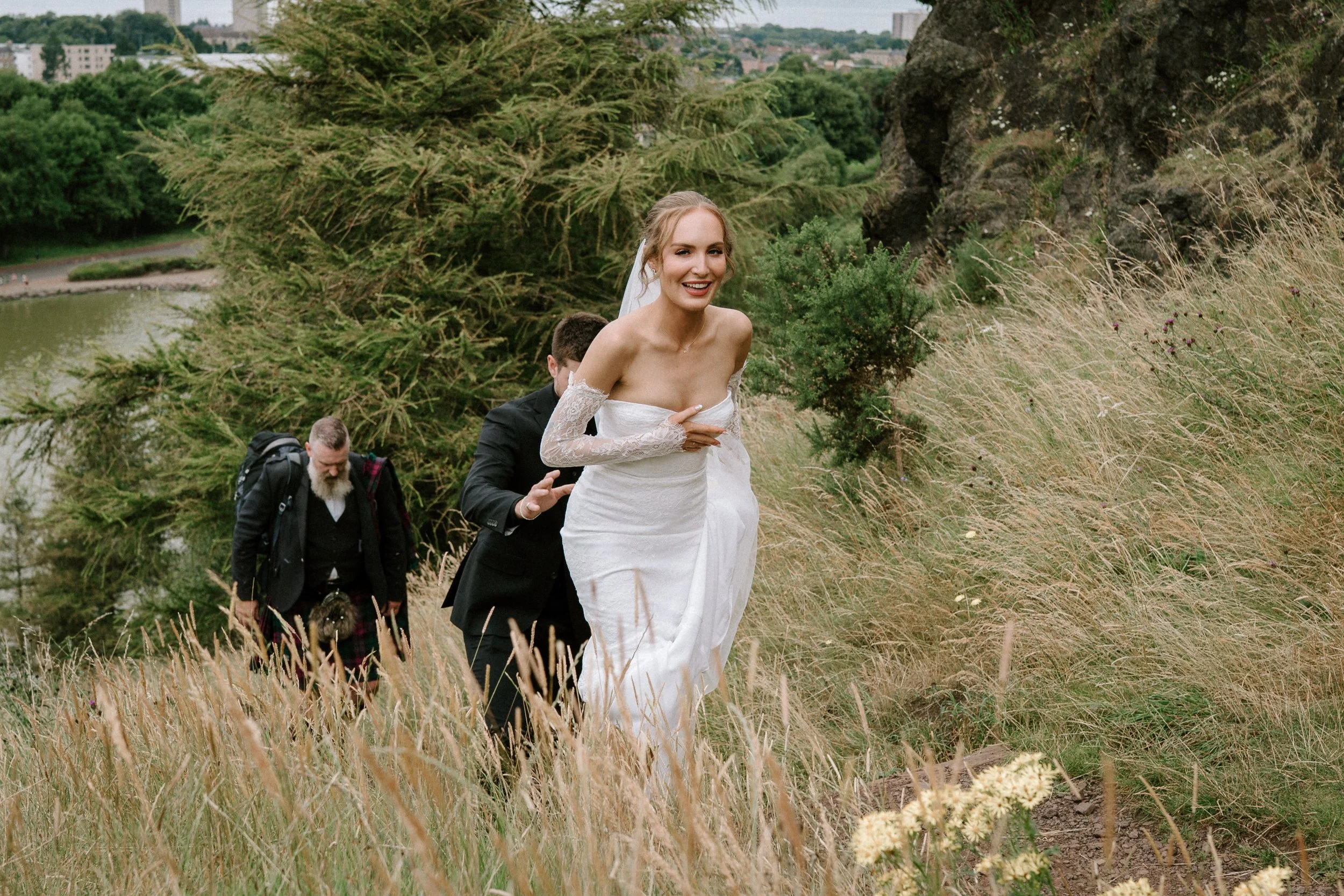 A bride in a white wedding dress and lace sleeves walking through tall grass in an outdoor area, smiling, with three men in formal attire walking behind her. - captured by an Edinburgh wedding photographer