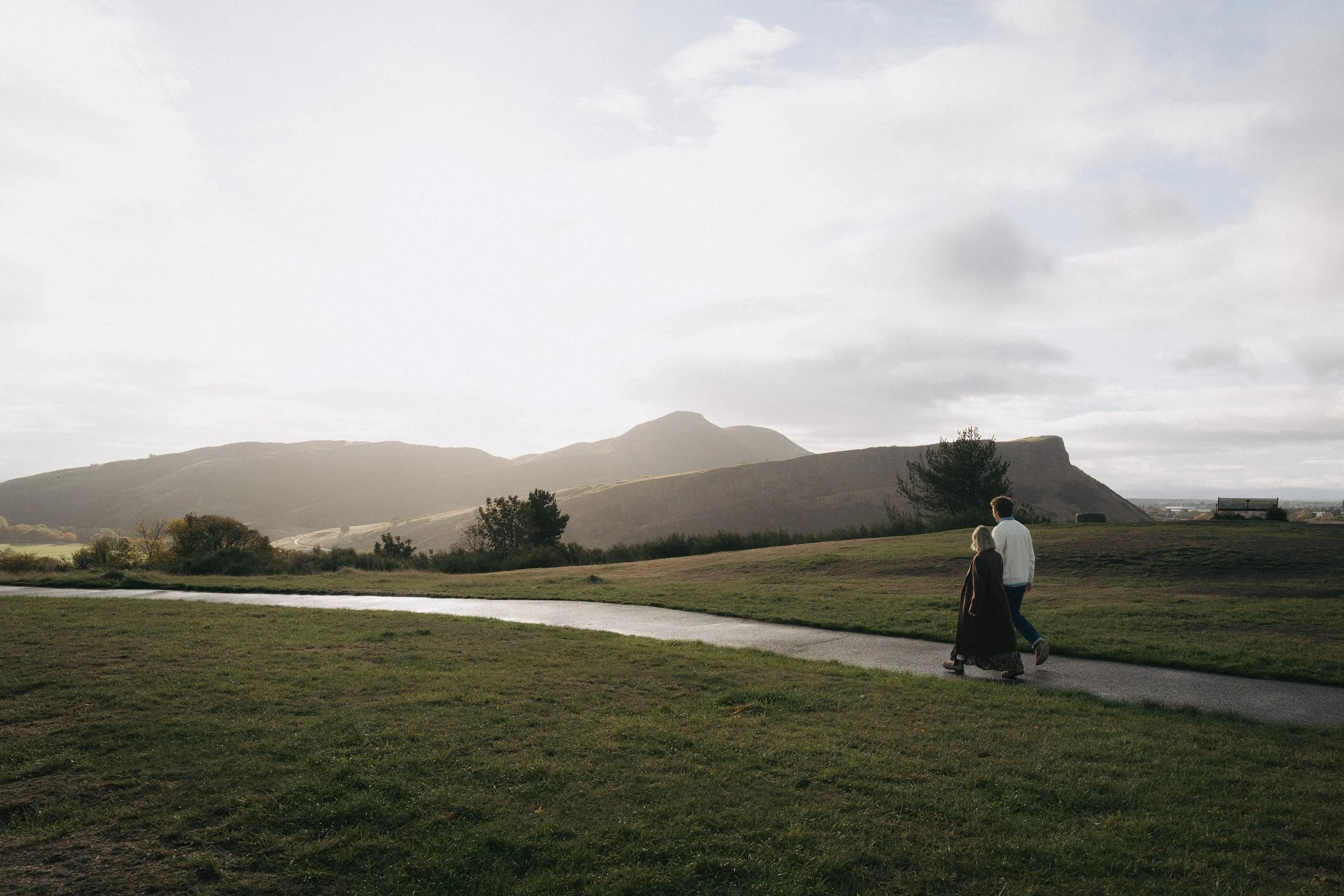 A man and woman walking on a winding pathway in a grassy park with hills and mountains in the background during overcast weather. - captured by an Edinburgh wedding photographer