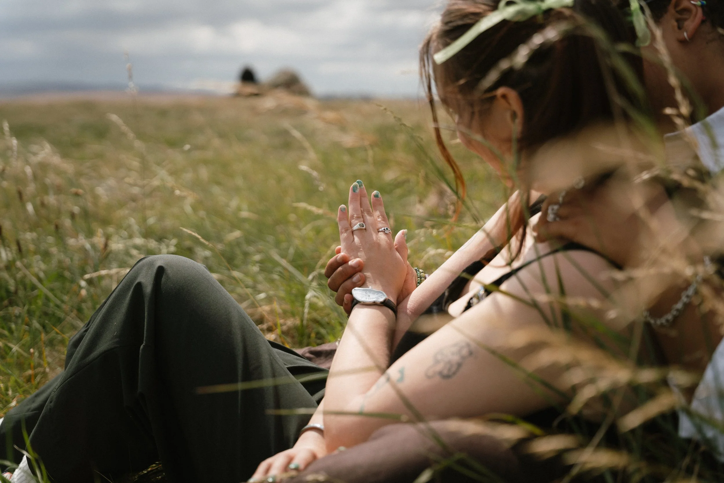 A woman with tattoos and rings, wearing a flower crown and jewelry, lying on grass in a field, holding hands with another person. - captured by an Edinburgh wedding photographer