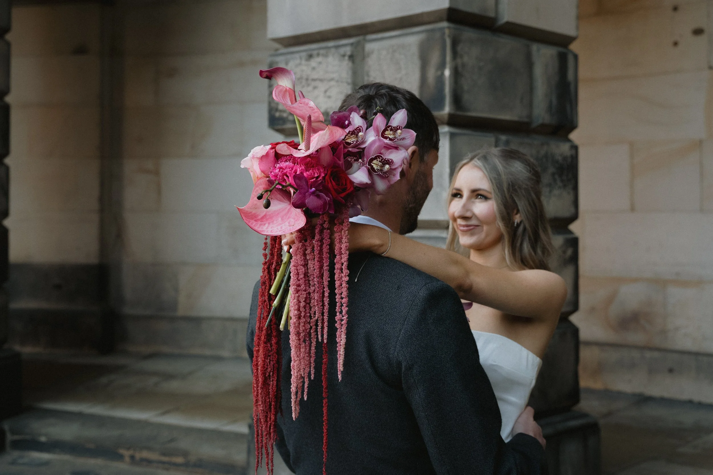 A woman and man embrace in a wedding setting, with the man holding a bouquet of pink, purple, and red flowers, and the woman smiling. - captured by an Edinburgh wedding photographer