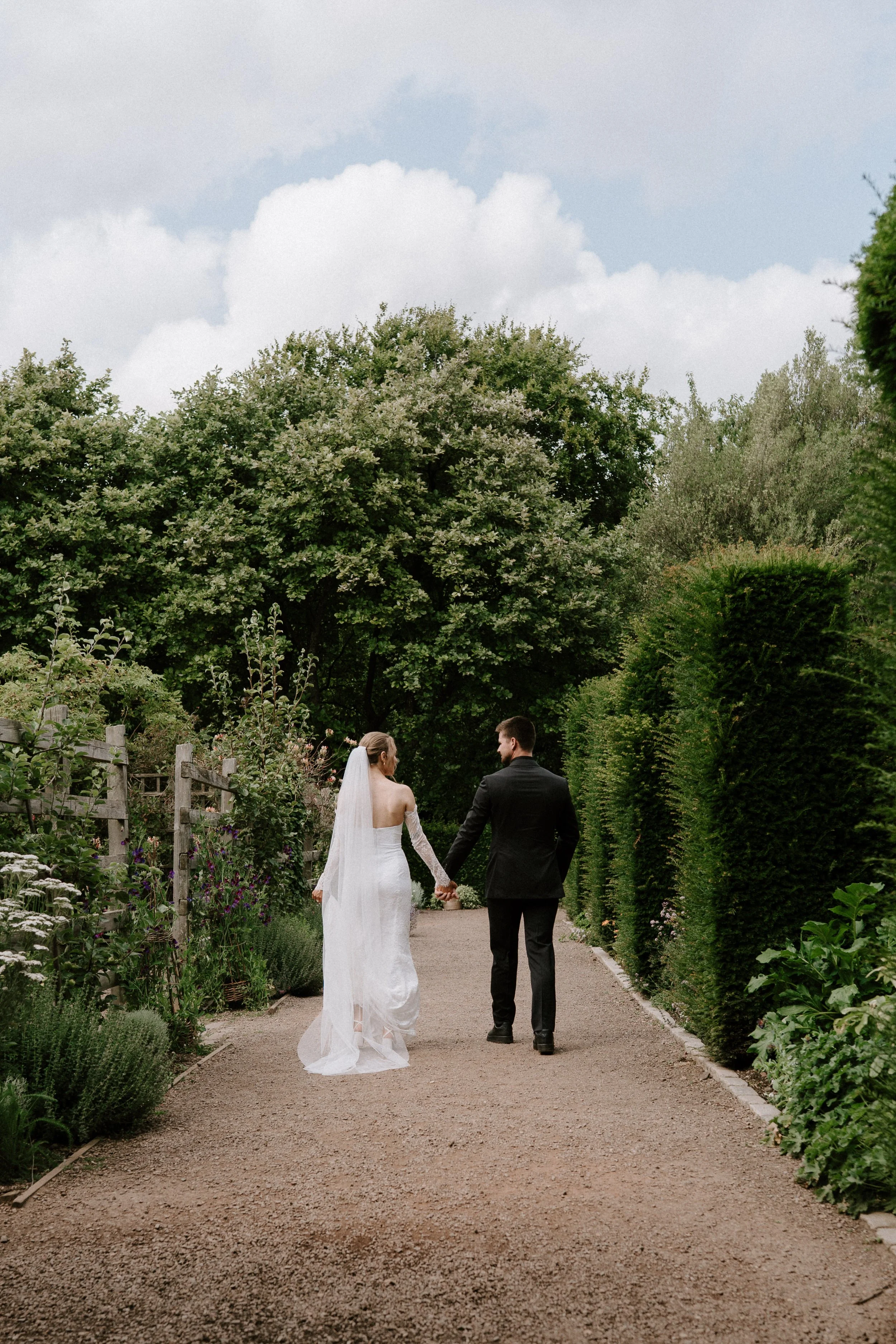 A bride and groom walking hand in hand through a garden pathway, surrounded by green trees and bushes, with a cloudy sky overhead. - captured by an Edinburgh wedding photographer