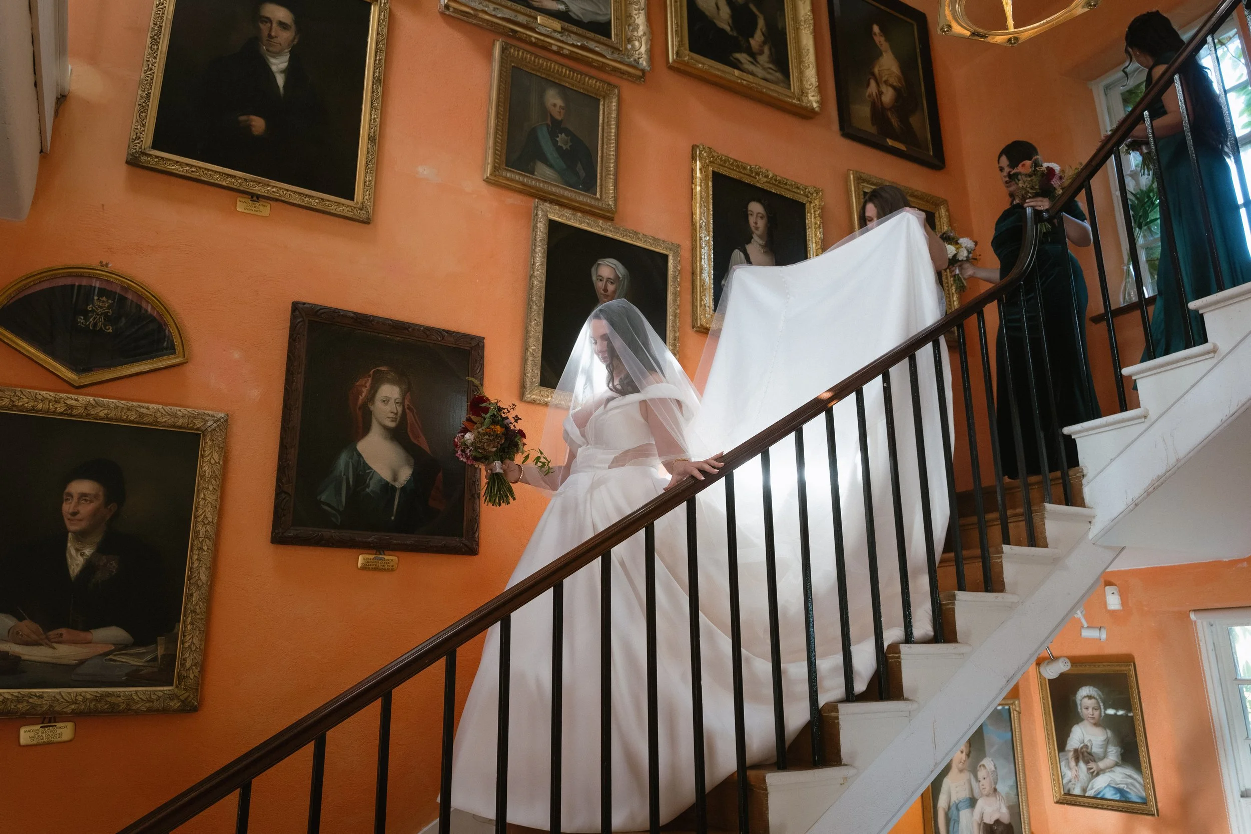 A bride in a white wedding gown with a veil and bouquet walking down a staircase in an art gallery with framed portraits on an orange wall. - captured by an Edinburgh wedding photographer